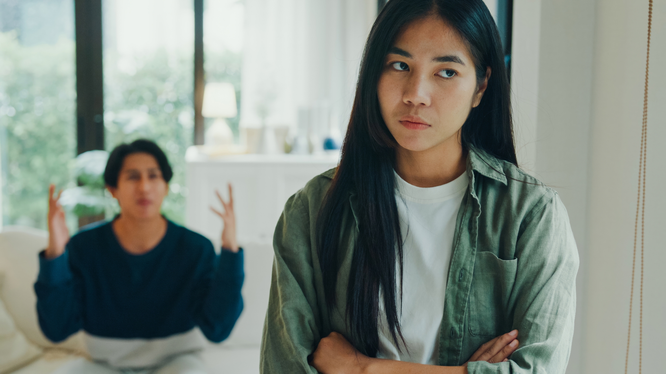 Woman looks away with arms crossed while person in background gestures in frustration in a room