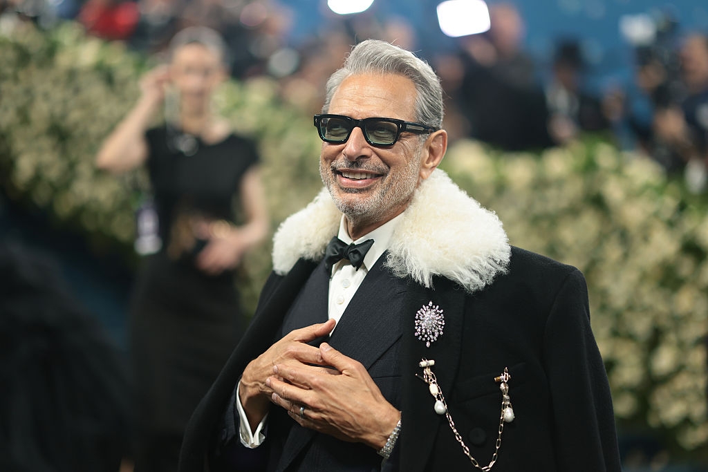 Man in tuxedo with fur-collared coat and brooch, smiling on a formal event carpet