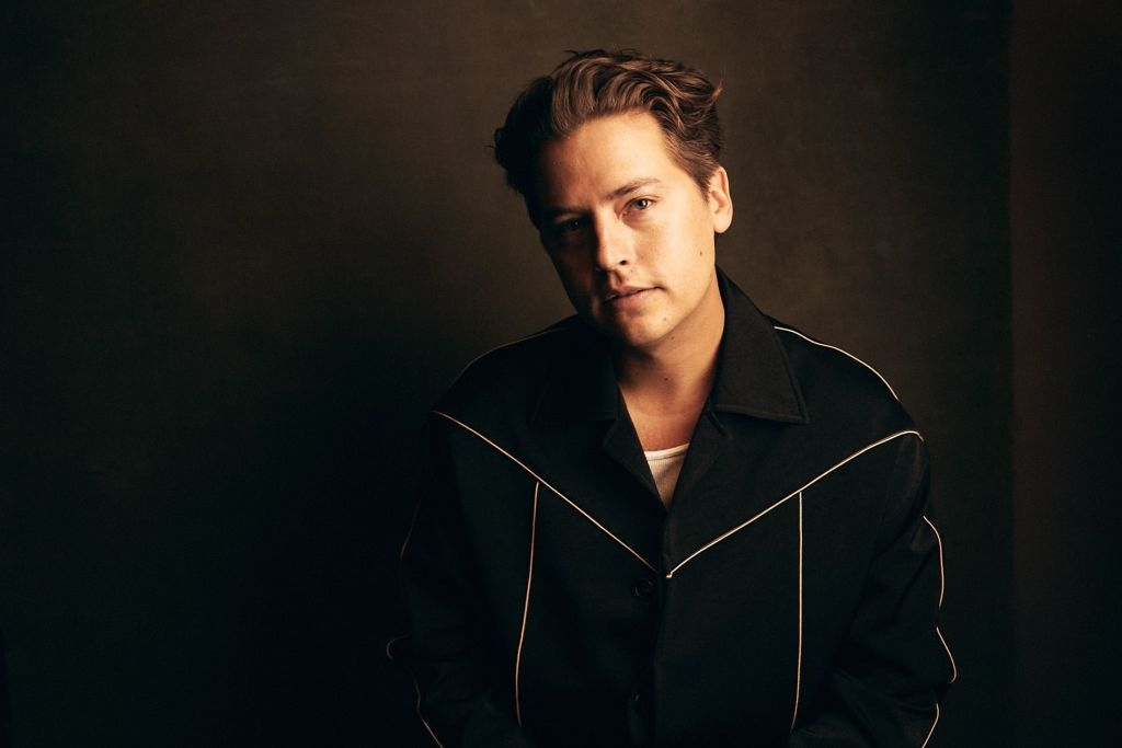 A person poses against a dark background, wearing a dark jacket with light piping details and a white shirt underneath, looking thoughtful