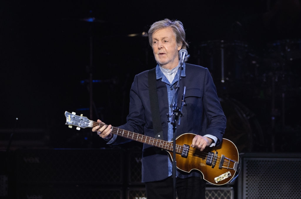 Musician performing on stage, holding a bass guitar, wearing a dark suit jacket over a light shirt