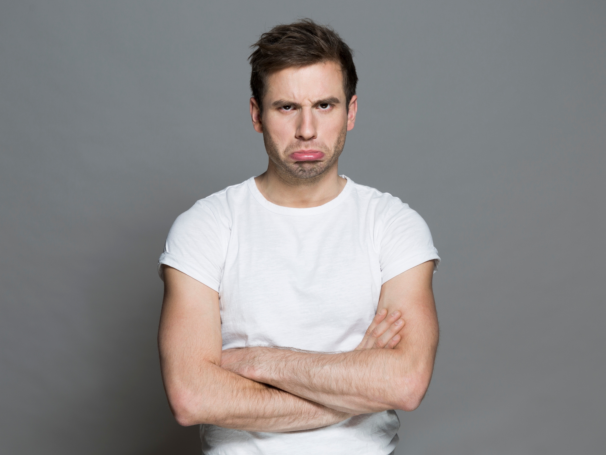 Man in a plain t-shirt with a pouting expression and crossed arms, looking directly at the camera against a plain background