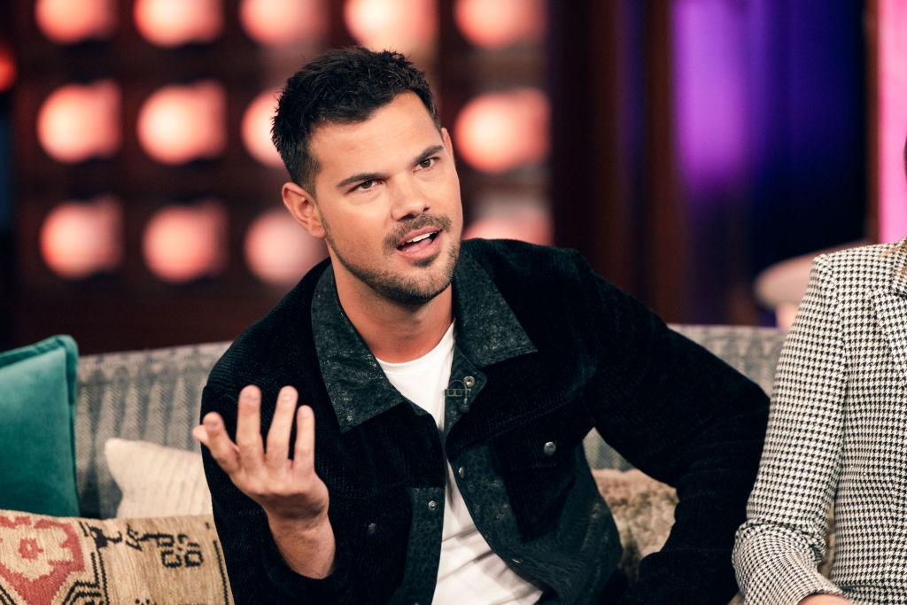 A person sits on a talk show couch wearing a casual jacket and white shirt, gesturing with one hand while speaking