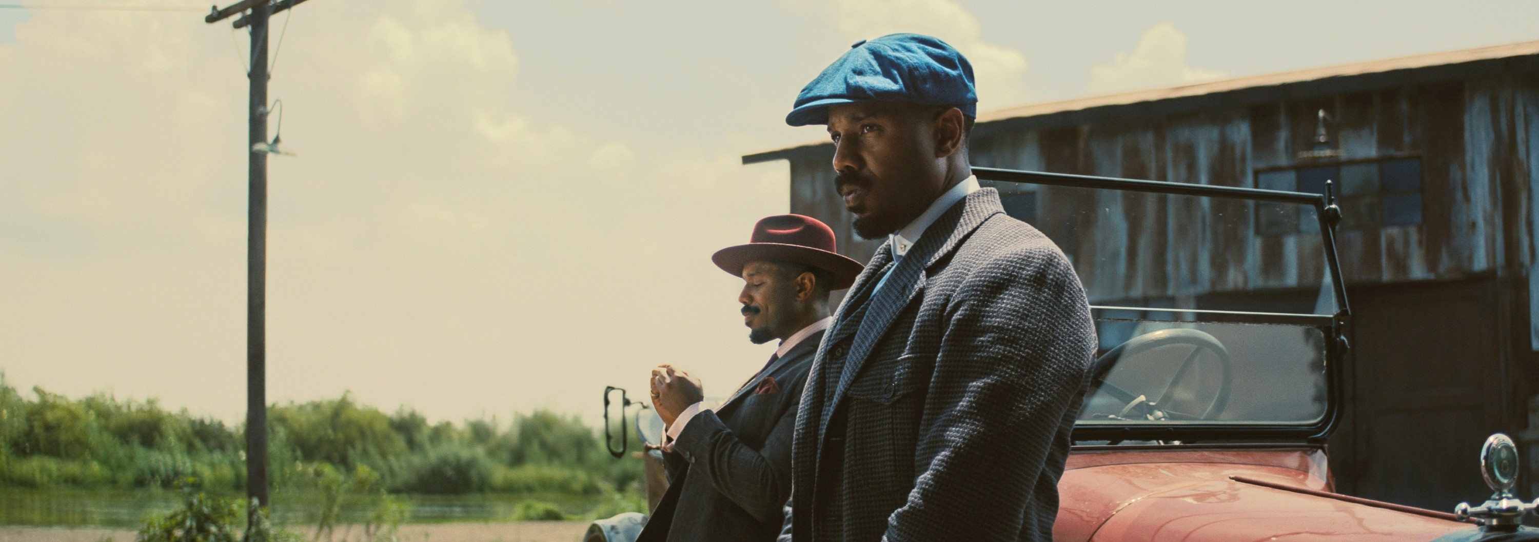Two men in vintage suits and hats stand beside a classic car, evoking a historical setting