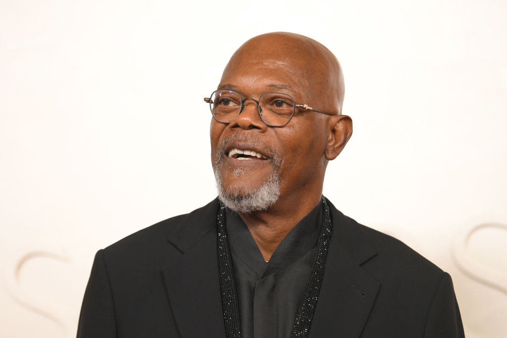 A person wearing a sleek black suit and glasses stands against a plain background, smiling slightly
