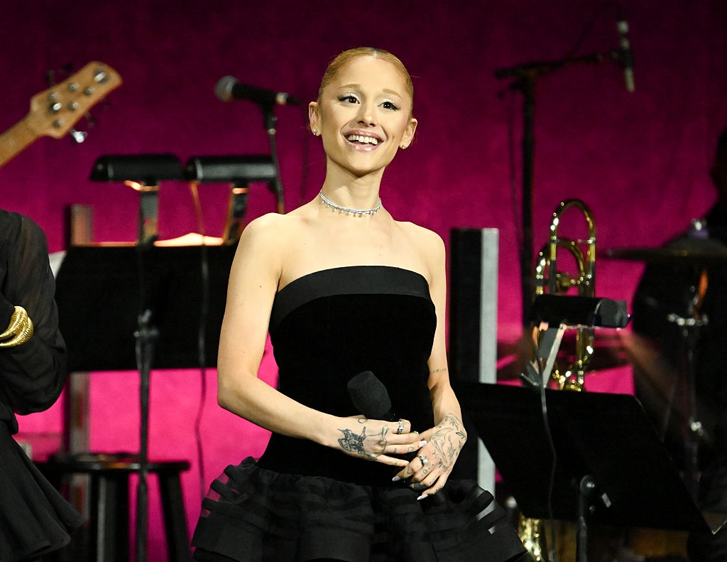 Person on stage in a strapless dress, holding a microphone, smiling with musical instruments in the background