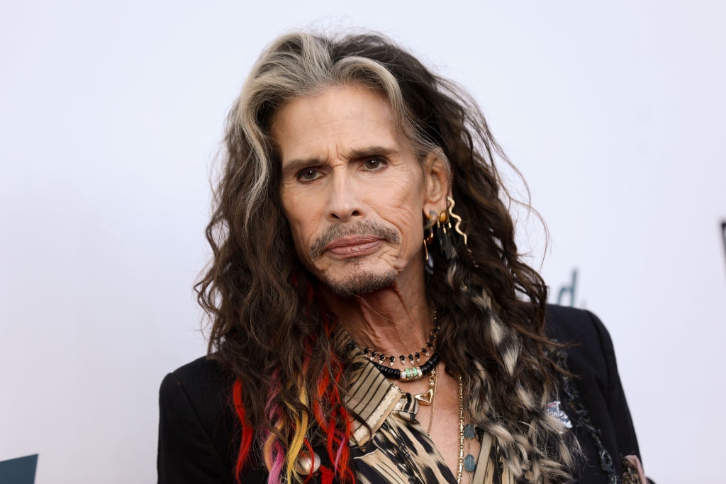 A person with long, wavy hair, wearing layered necklaces and a patterned shirt, photographed at a celebrity event