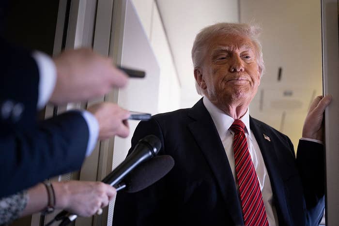 A person in a suit and tie stands in a hallway while reporters hold out microphones and recording devices