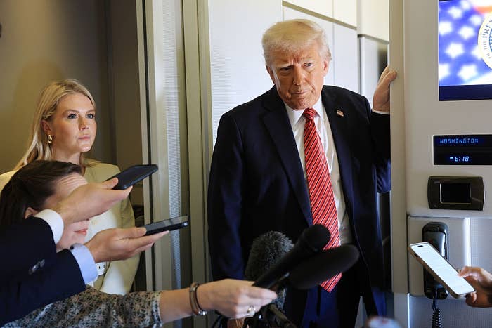 A man in a suit addresses reporters on an airplane, with microphones and phones directed towards him