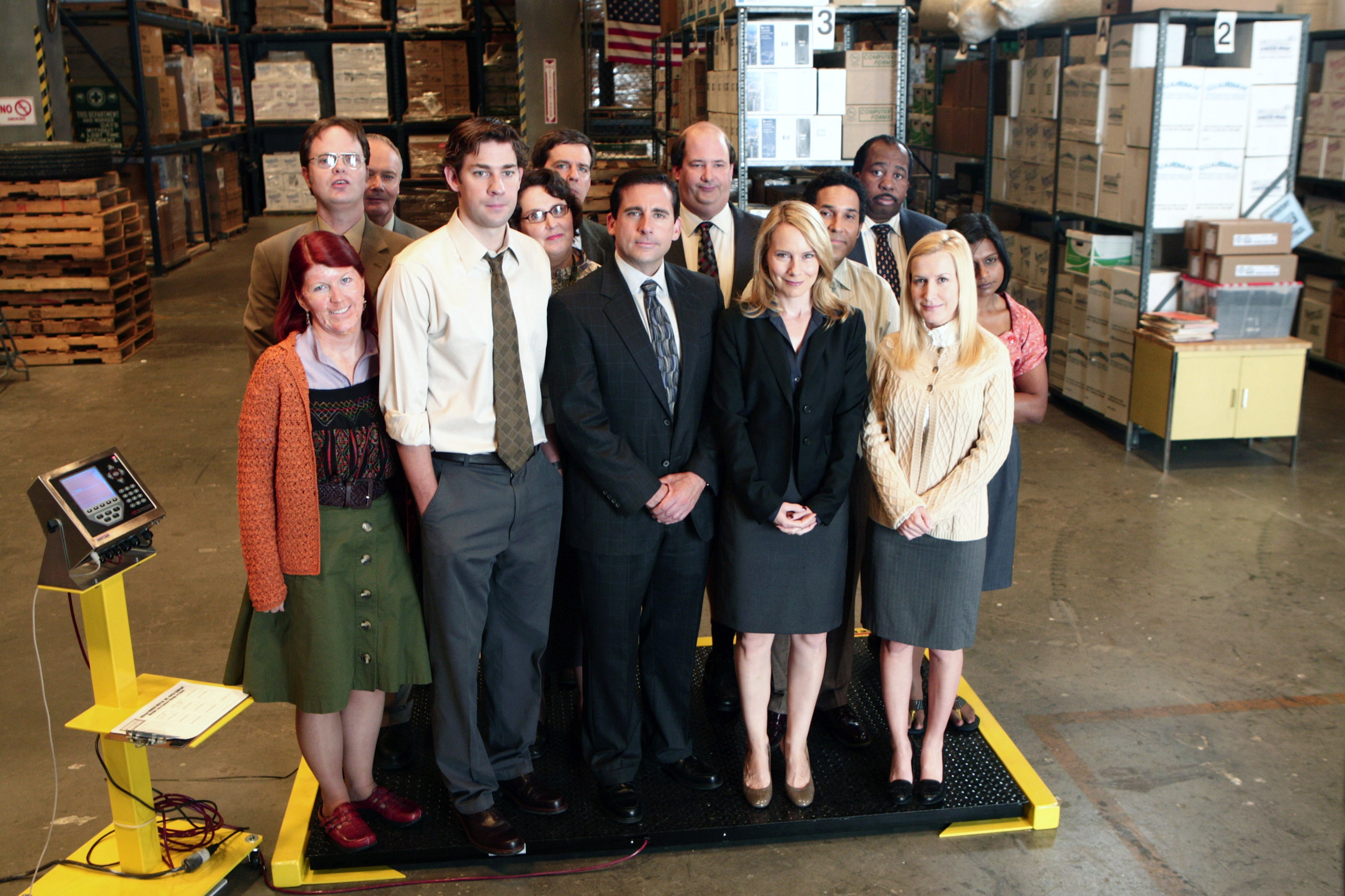 Cast of a TV show posing in a warehouse setting; actors wear business and casual attire, including suits, a cardigan, and a skirt