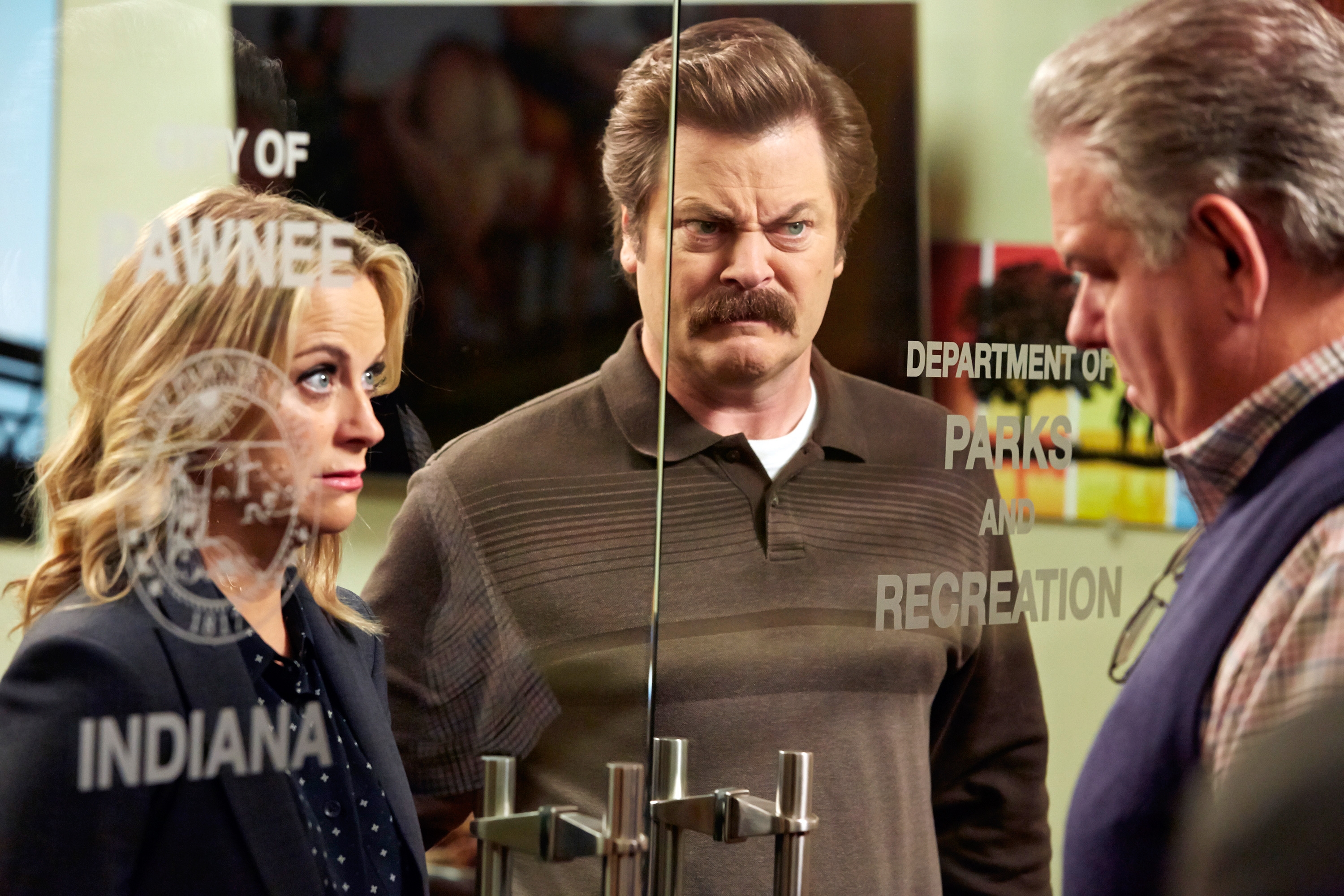 Two people look through a glass door labeled &quot;Department of Parks and Recreation&quot; at a man inside an office. One person wears a blazer, the other a polo shirt