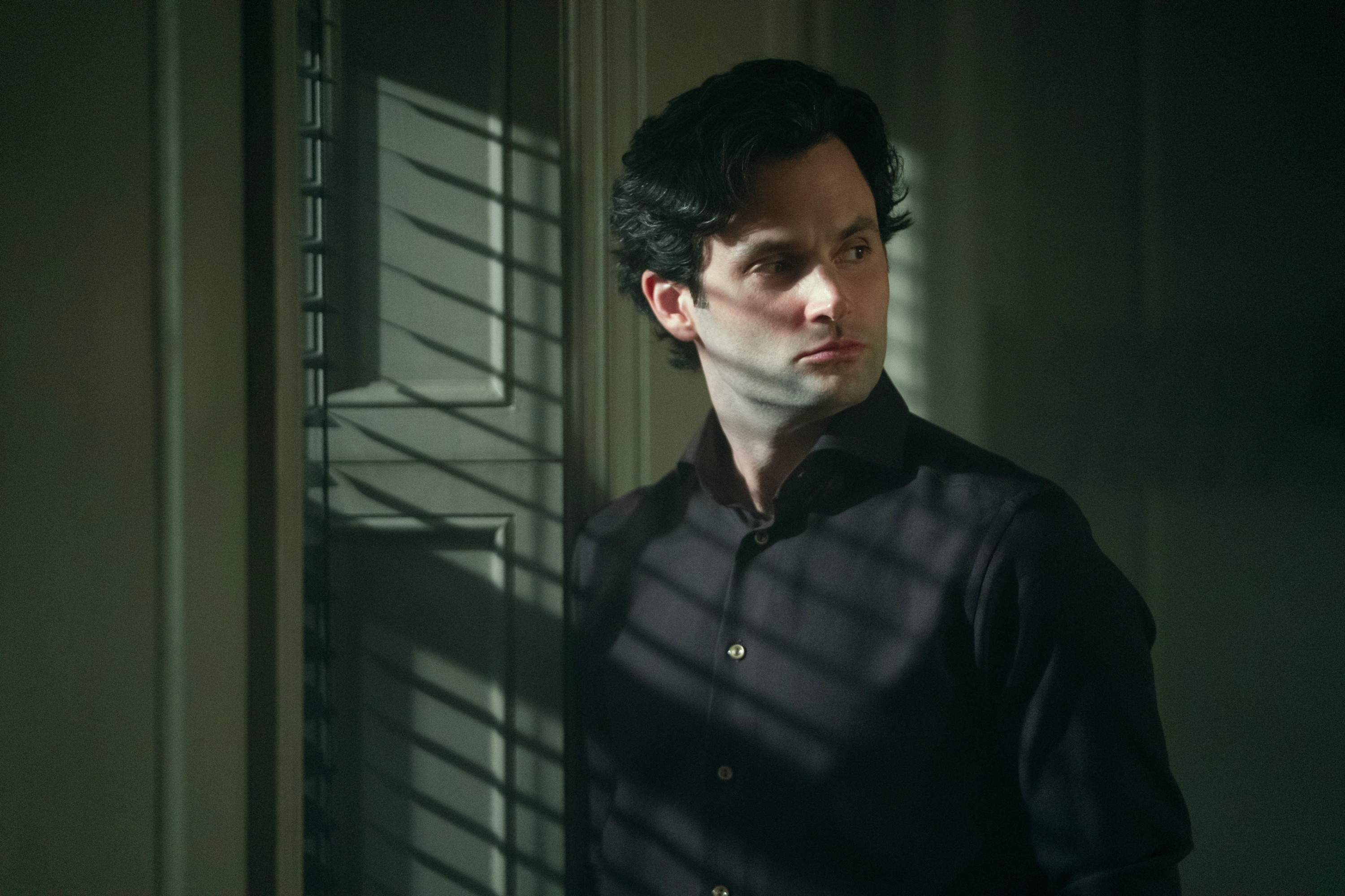 A man stands in front of window blinds, casting shadows on his serious expression, wearing a dark shirt in a dramatic scene