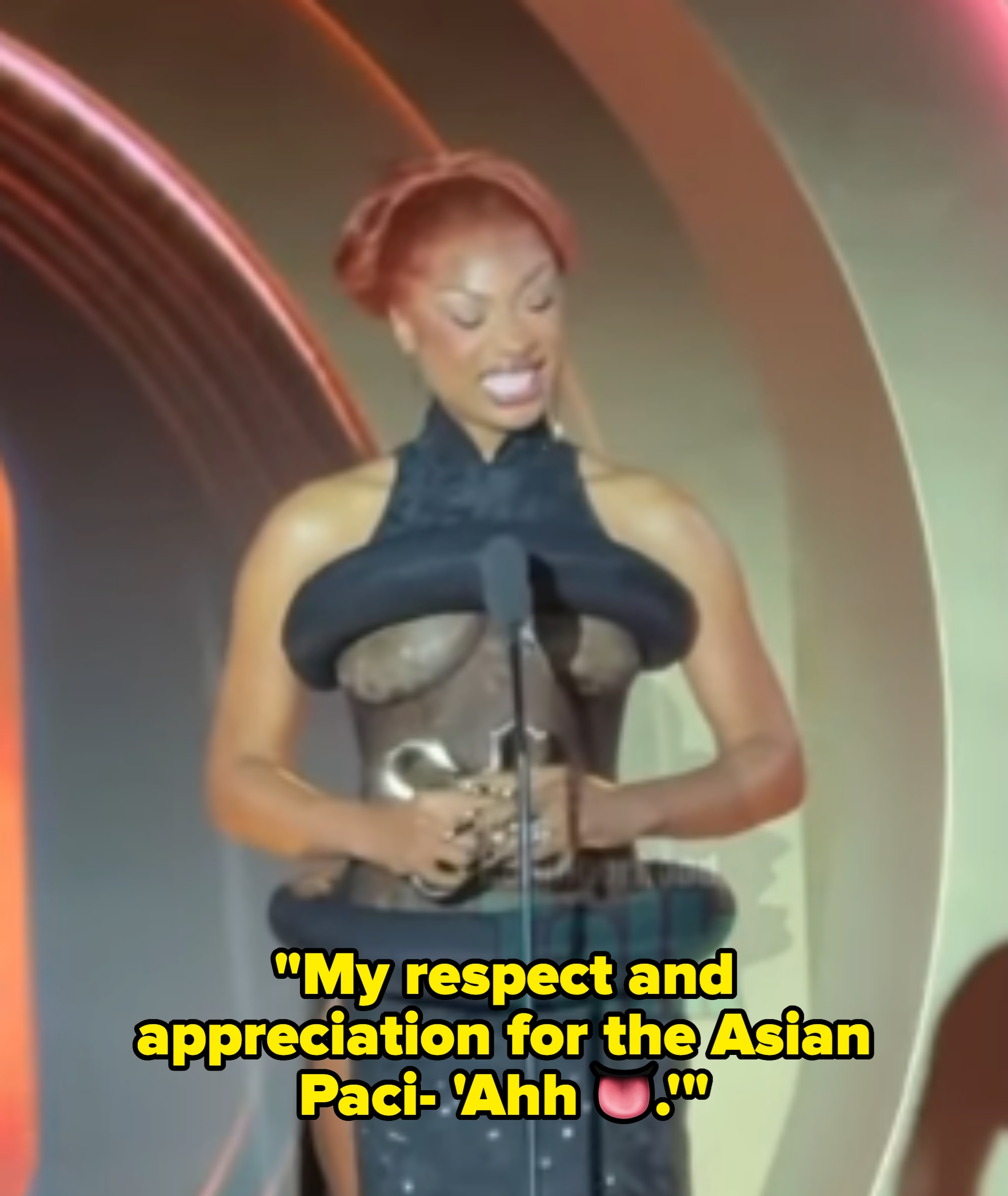 Person in a stylish, avant-garde outfit with a microphone in front, standing on stage at a formal event