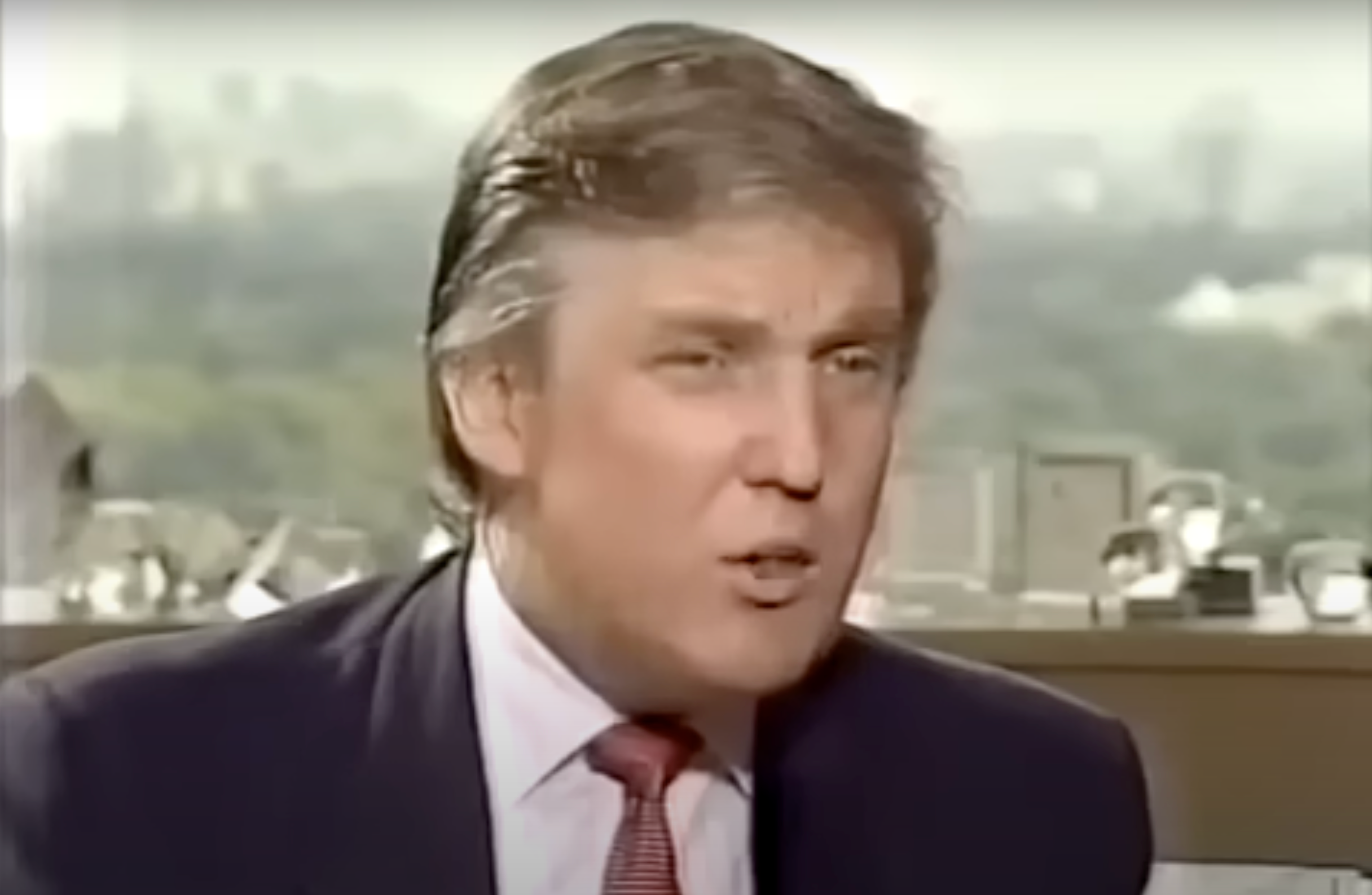 A person in a suit and tie is sitting indoors, appearing to speak during an interview