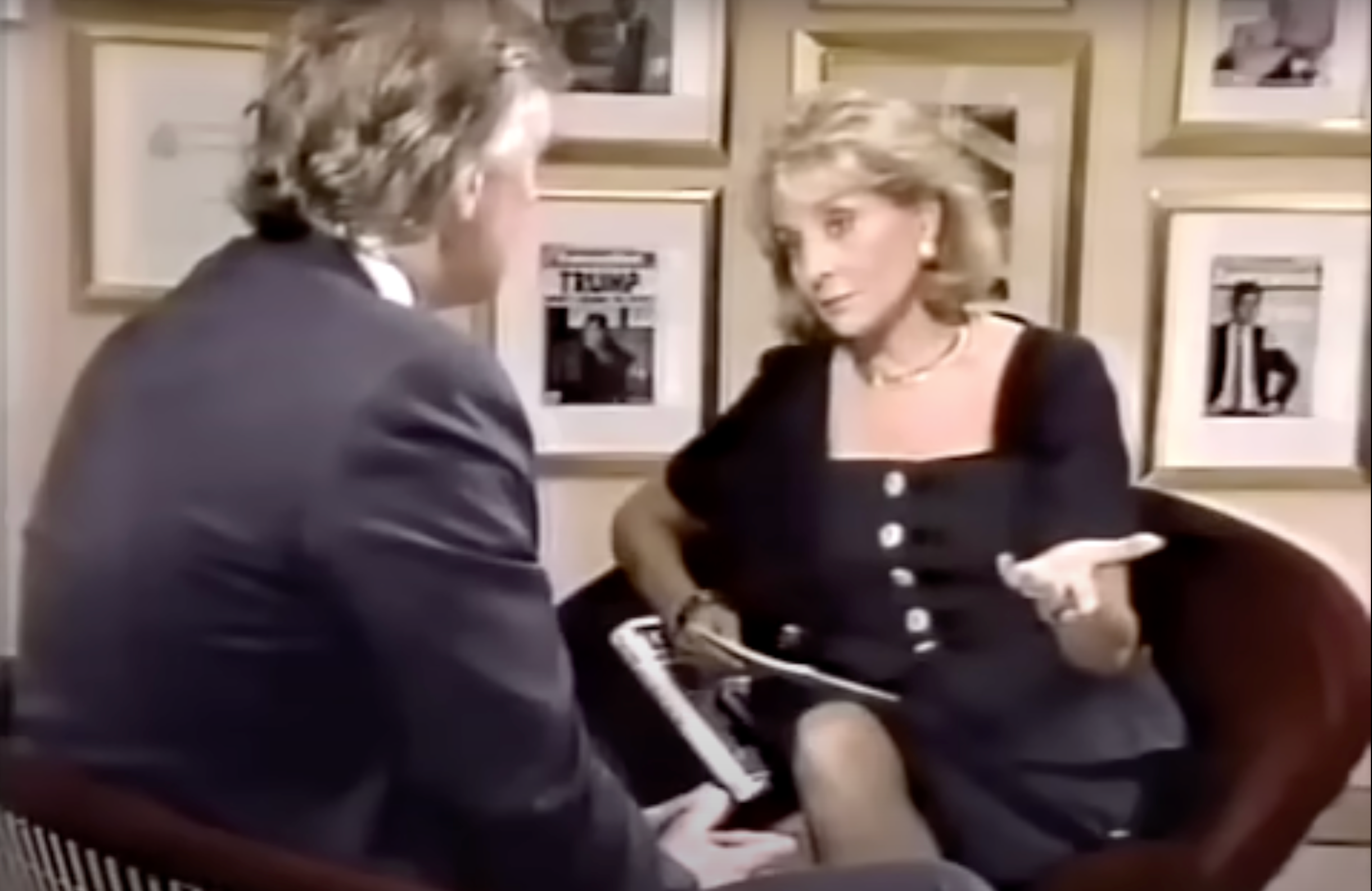 Two people sitting in an interview setting. The woman wears a classic tailored dress and holds papers, gesturing as she speaks to the man