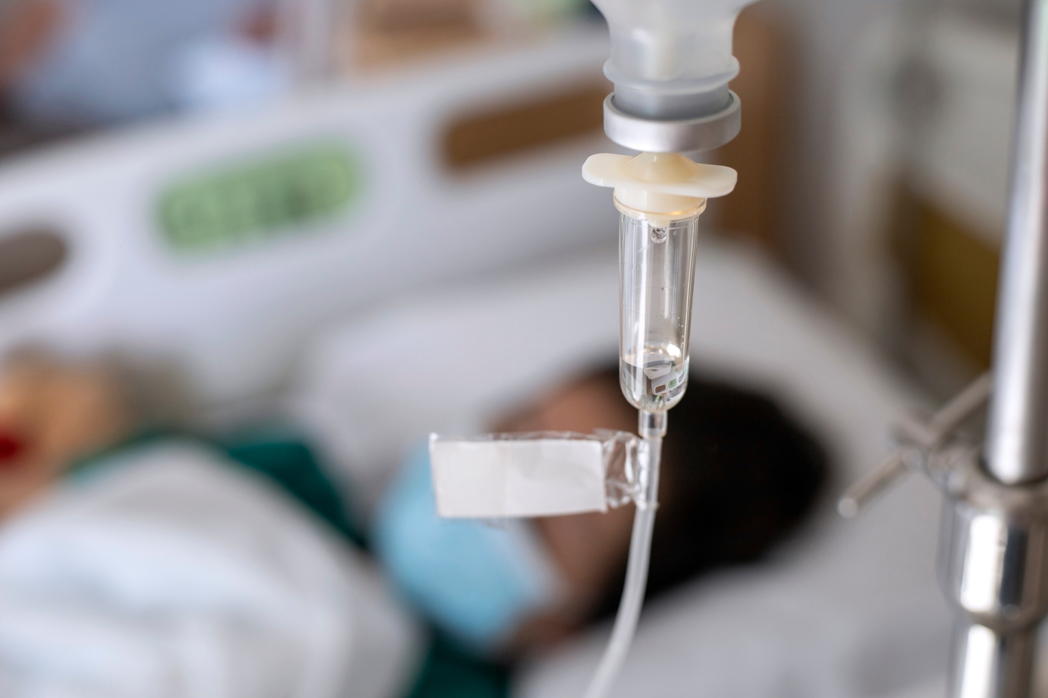 A person in a hospital bed with an IV line in focus, wearing a mask