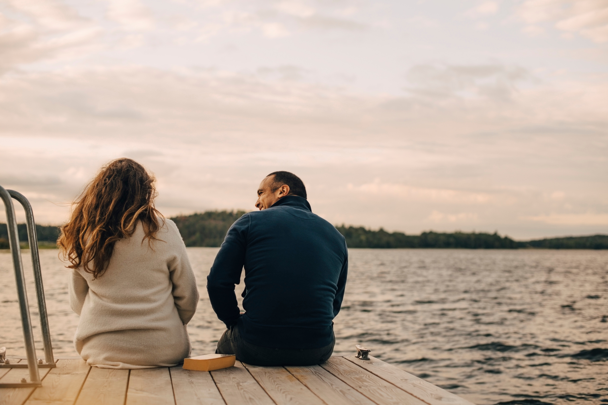 Two people sitting on a dock by a lake, engaged in conversation, with a serene background of water and distant trees