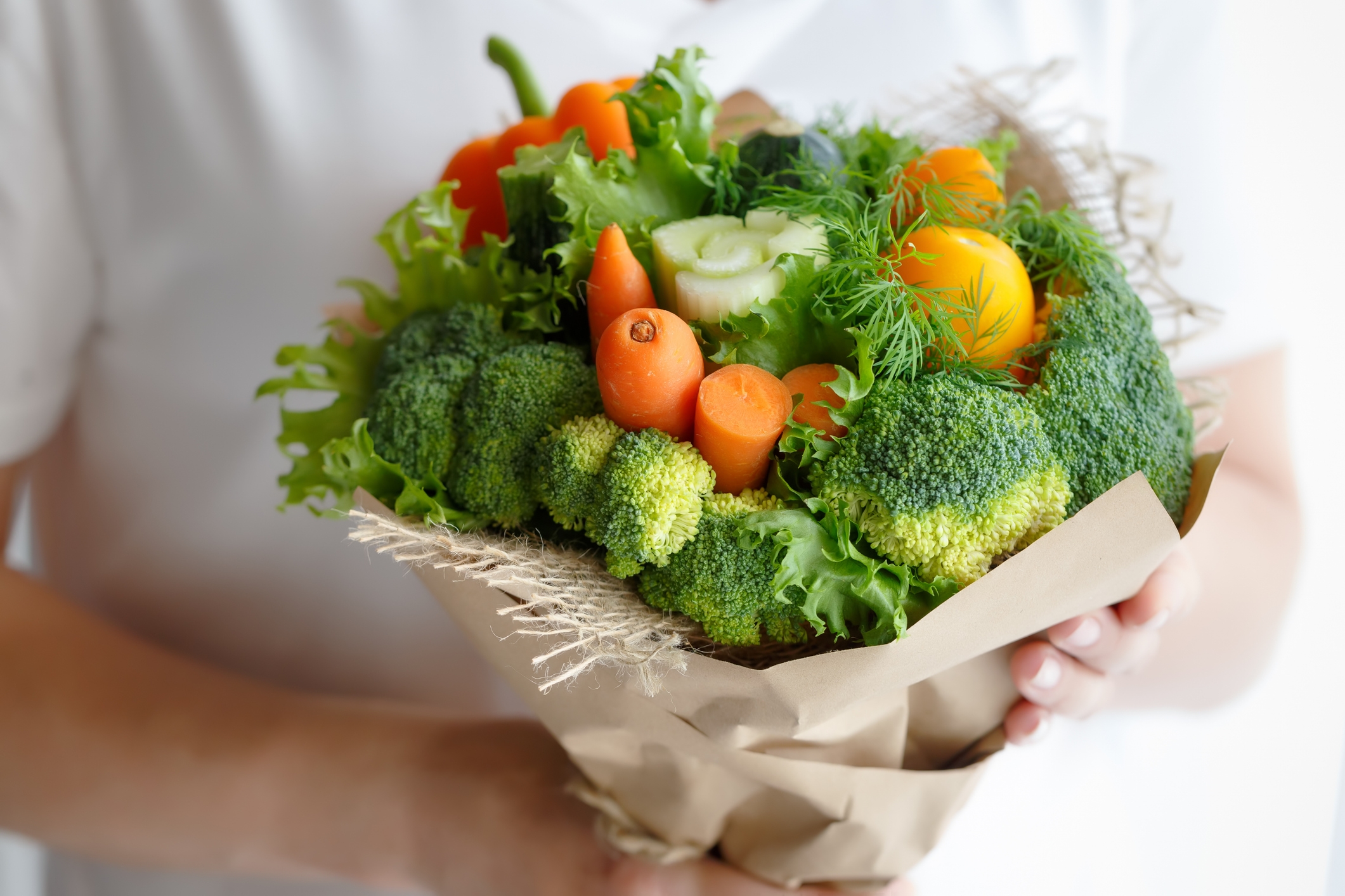 A person holds a bouquet made of assorted fresh vegetables like broccoli, carrots, and lettuce, wrapped in brown paper