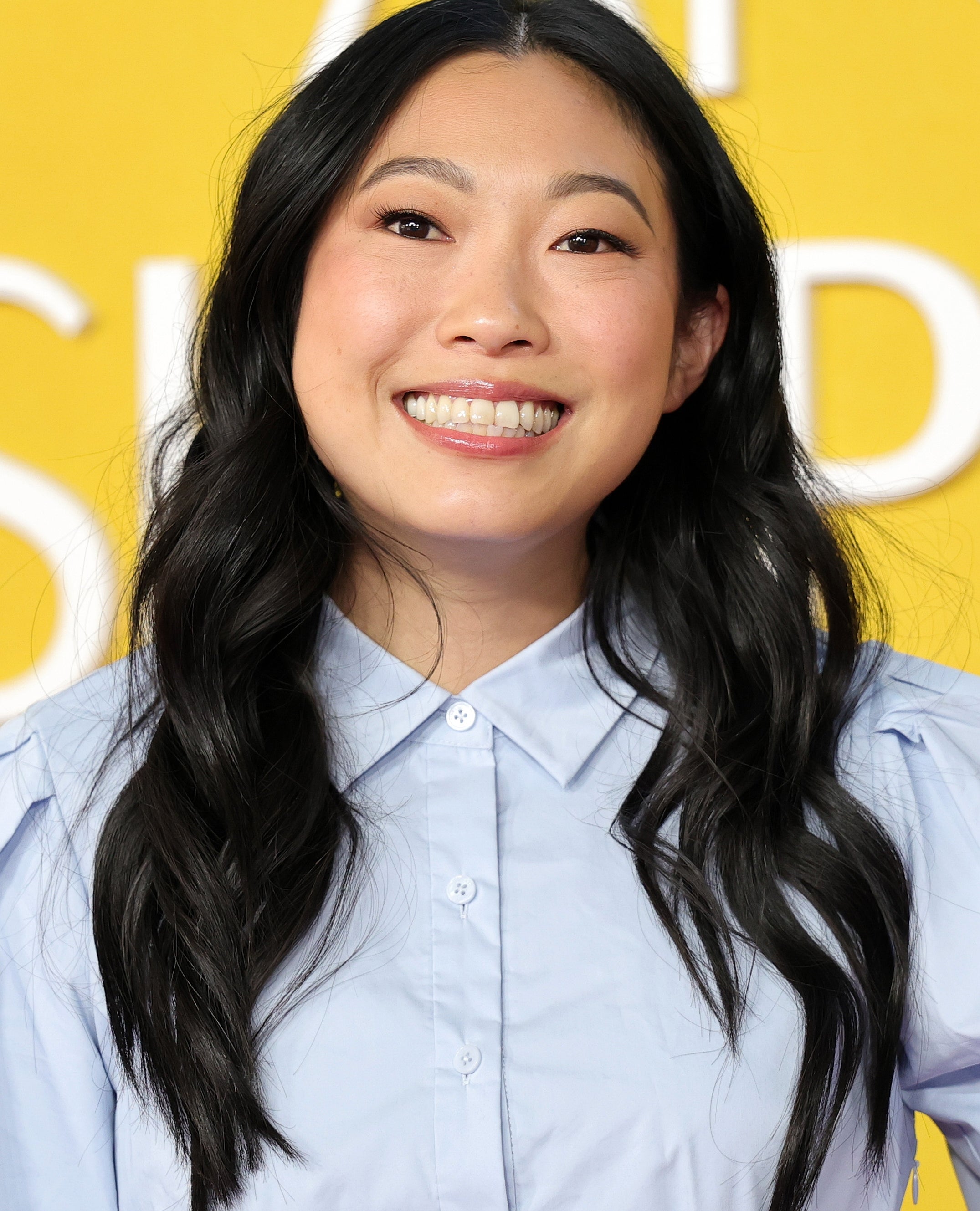 Awkwafina smiling, wearing a light blouse, with long wavy hair, against a text-covered backdrop