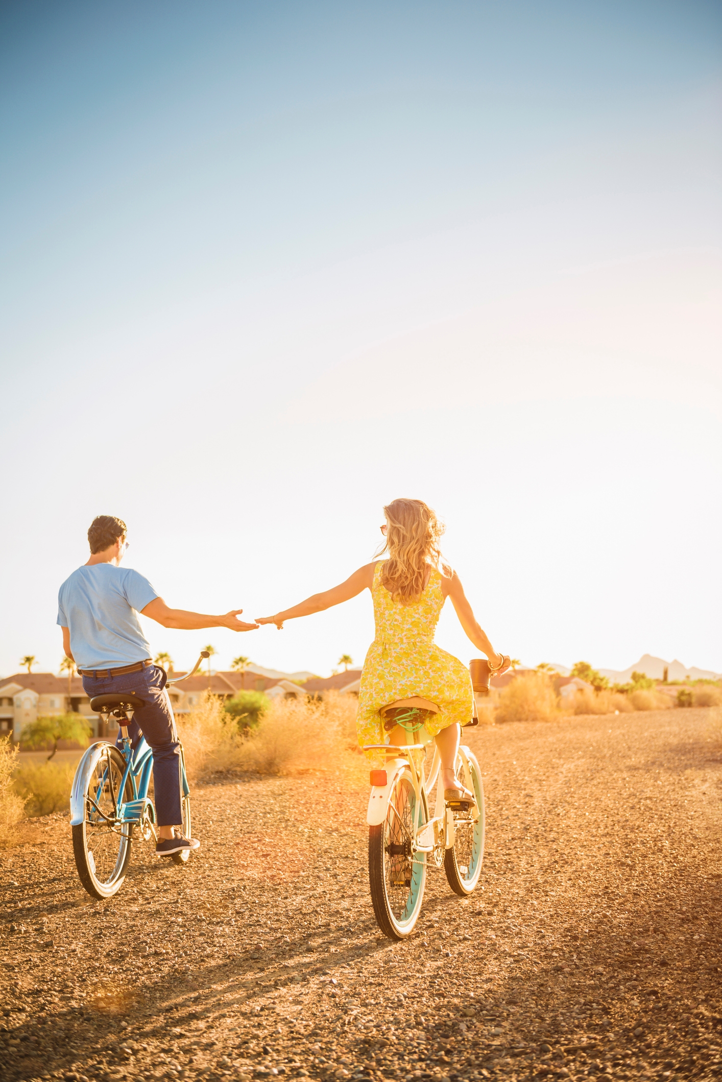 Two people ride bicycles on a gravel path, holding hands, with the sun setting in the background