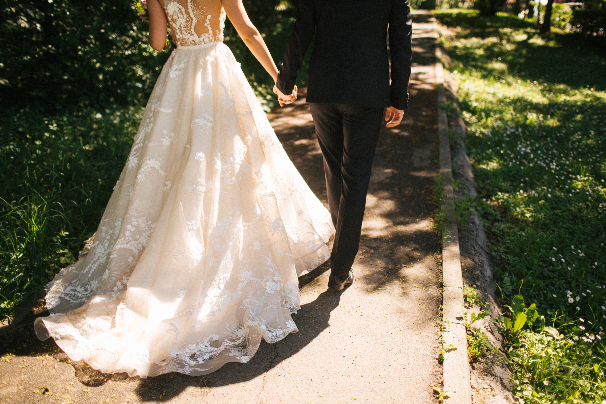 Couple in elegant wedding attire walking hand in hand down a sunlit path in a park