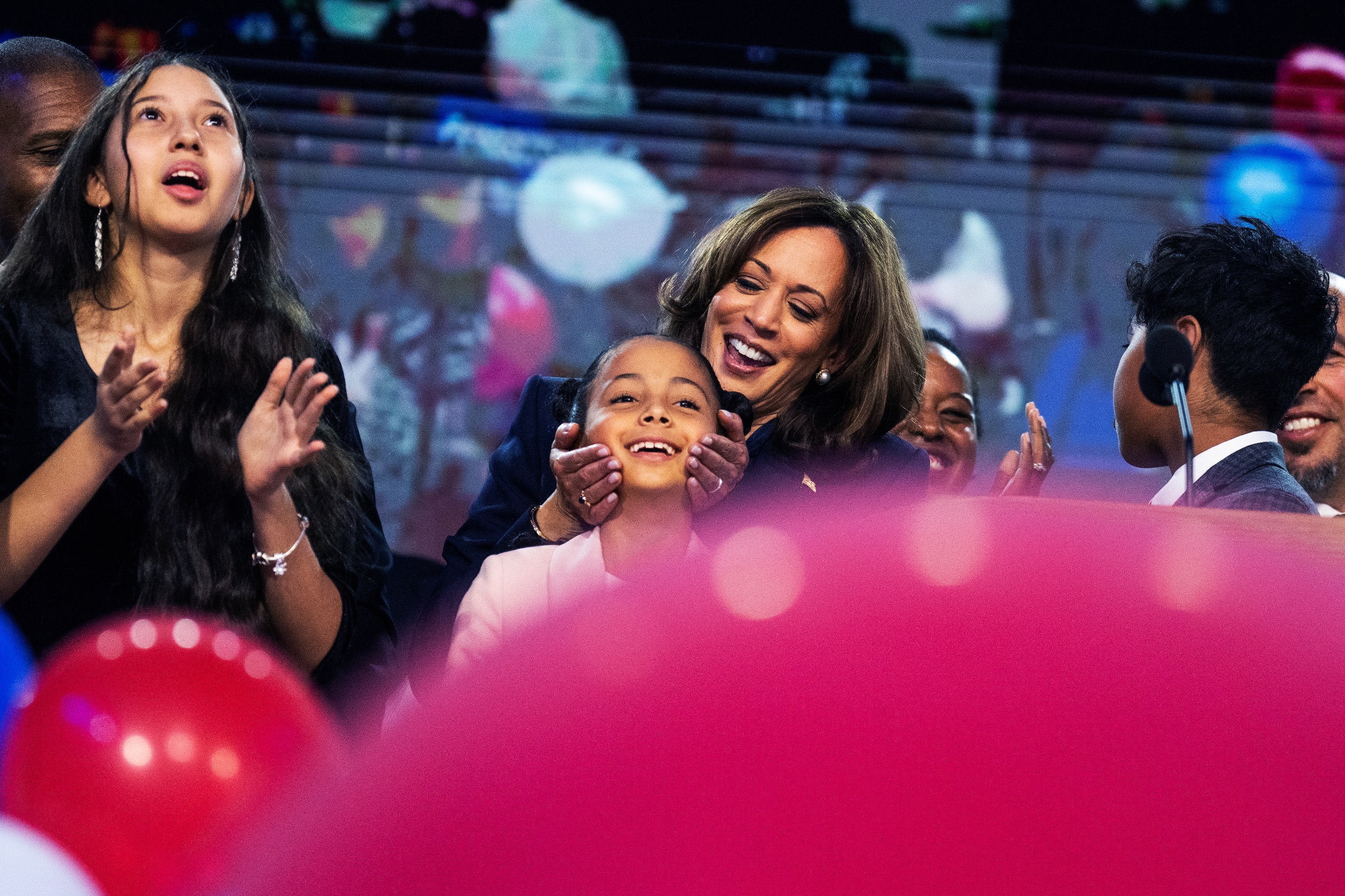 Group of people celebrating at an event with balloons, including a Kamala Harris embracing a child, all looking up joyfully