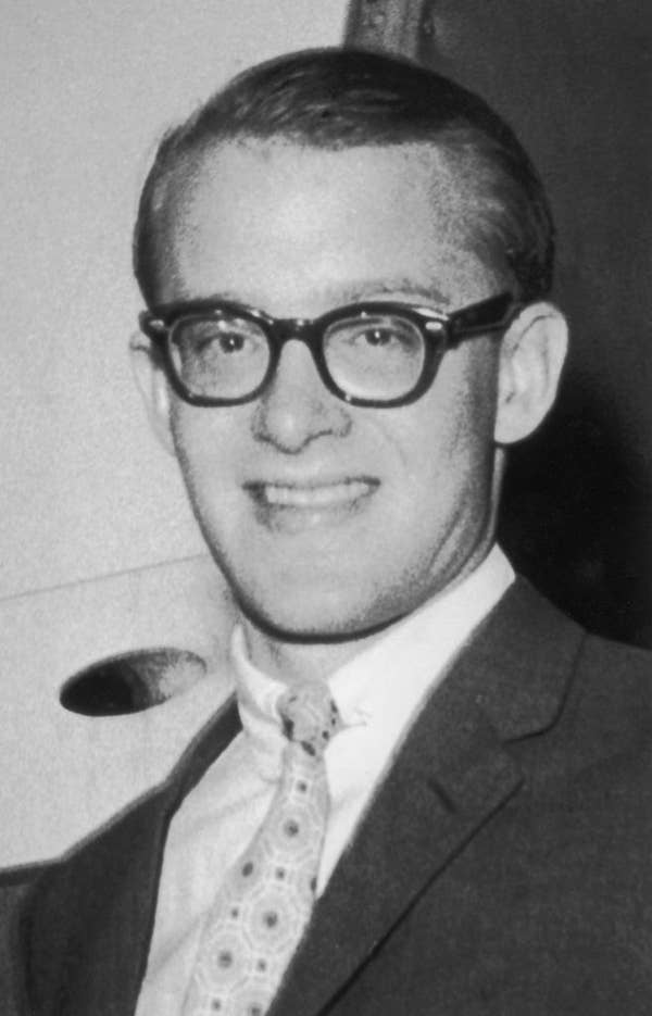 A young man in a suit, tie with geometric pattern, and glasses smiles warmly