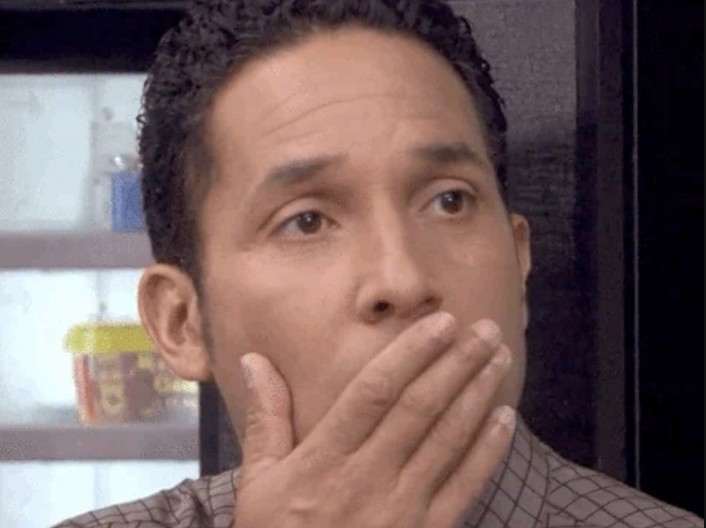 Oscar from The Office with a surprised expression, covering his mouth with a hand, suggesting shock or unexpected news