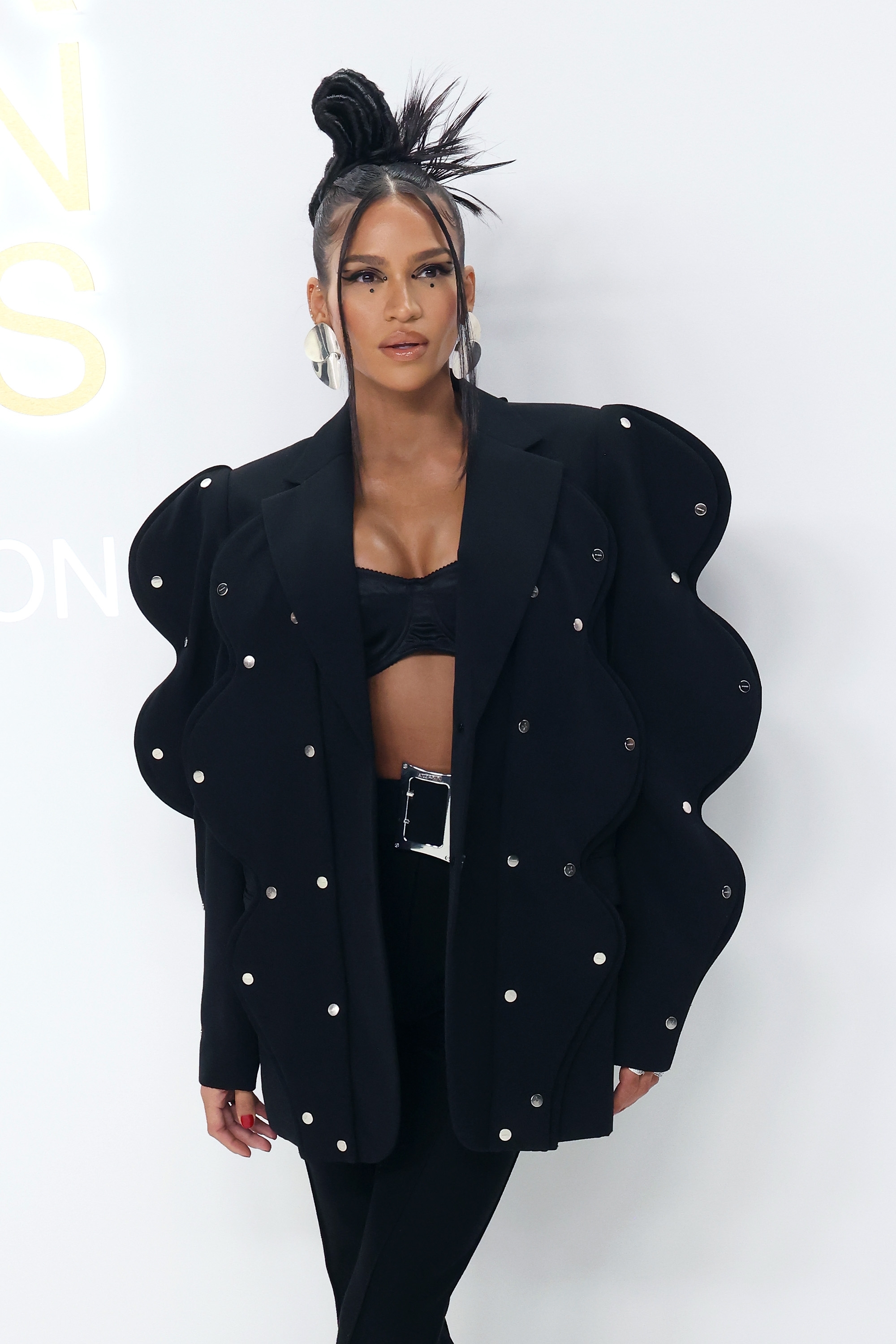 Person in avant-garde blazer with wave-like embellishments and crop top, posing at a fashion event. Hair styled in a high, artistic bun