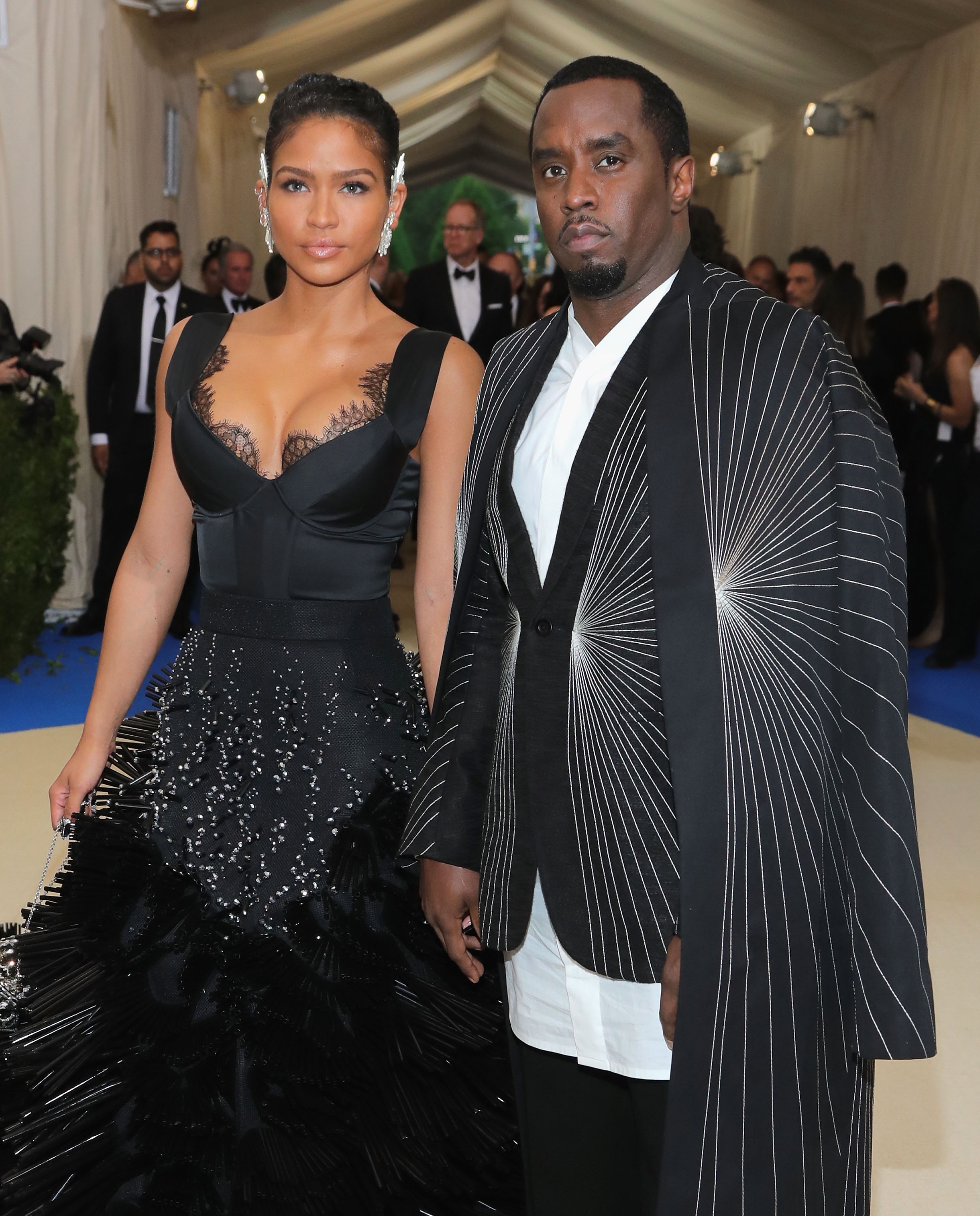 Two people on a formal event carpet. Woman in an elegant gown with lace details. Man in a structured suit with a geometric, radiating pattern cloak