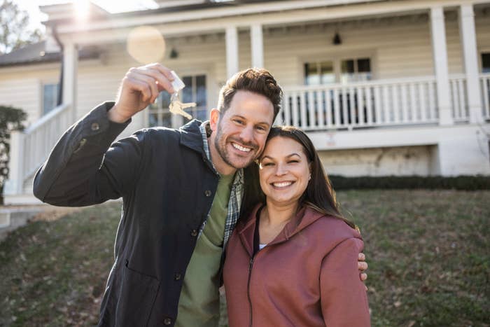 A smiling couple stands in front of a house, holding keys, symbolizing a new home purchase or real estate success