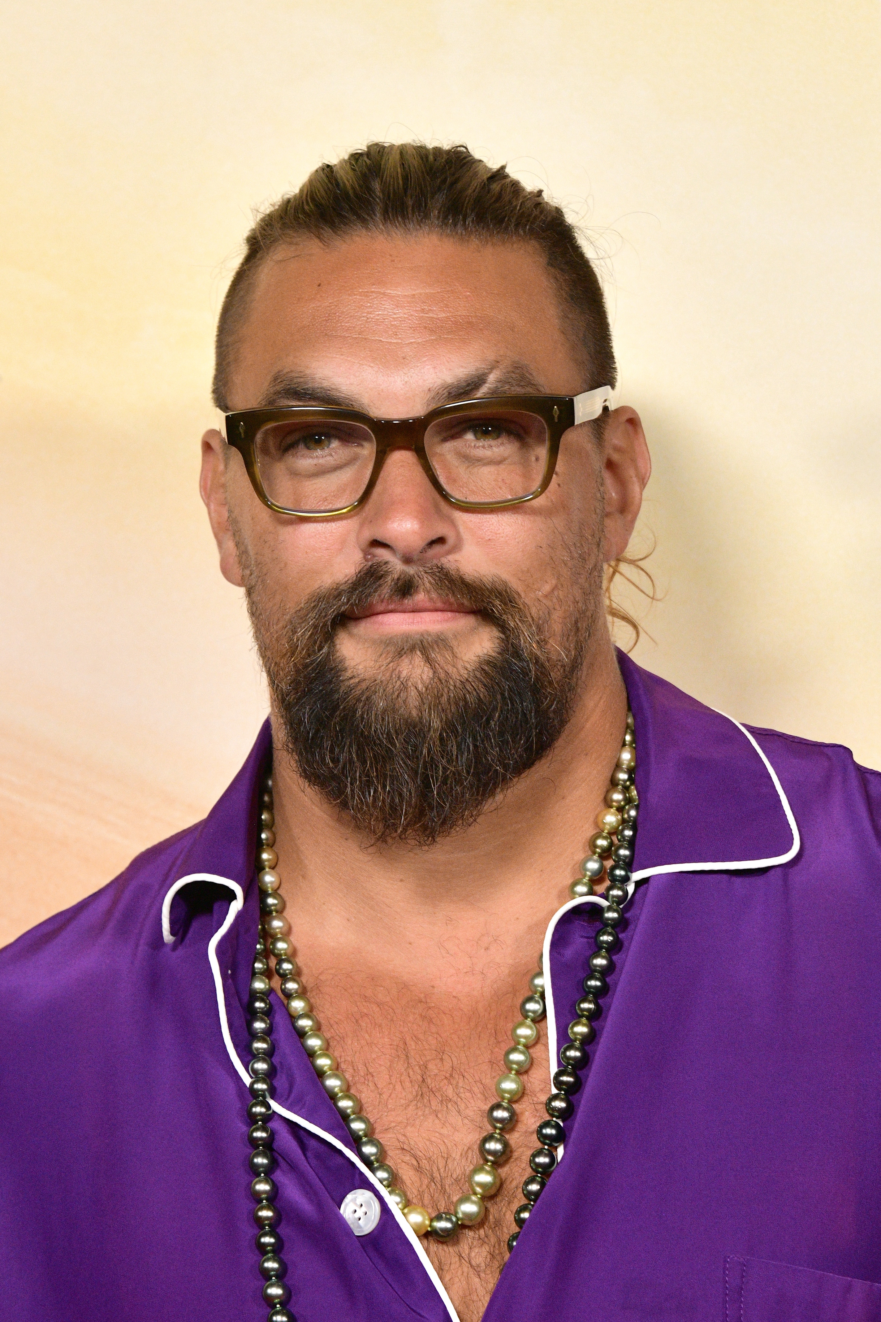 Jason Momoa with tied-back hair, glasses, and beard wearing a purple shirt with white trim and layered necklaces