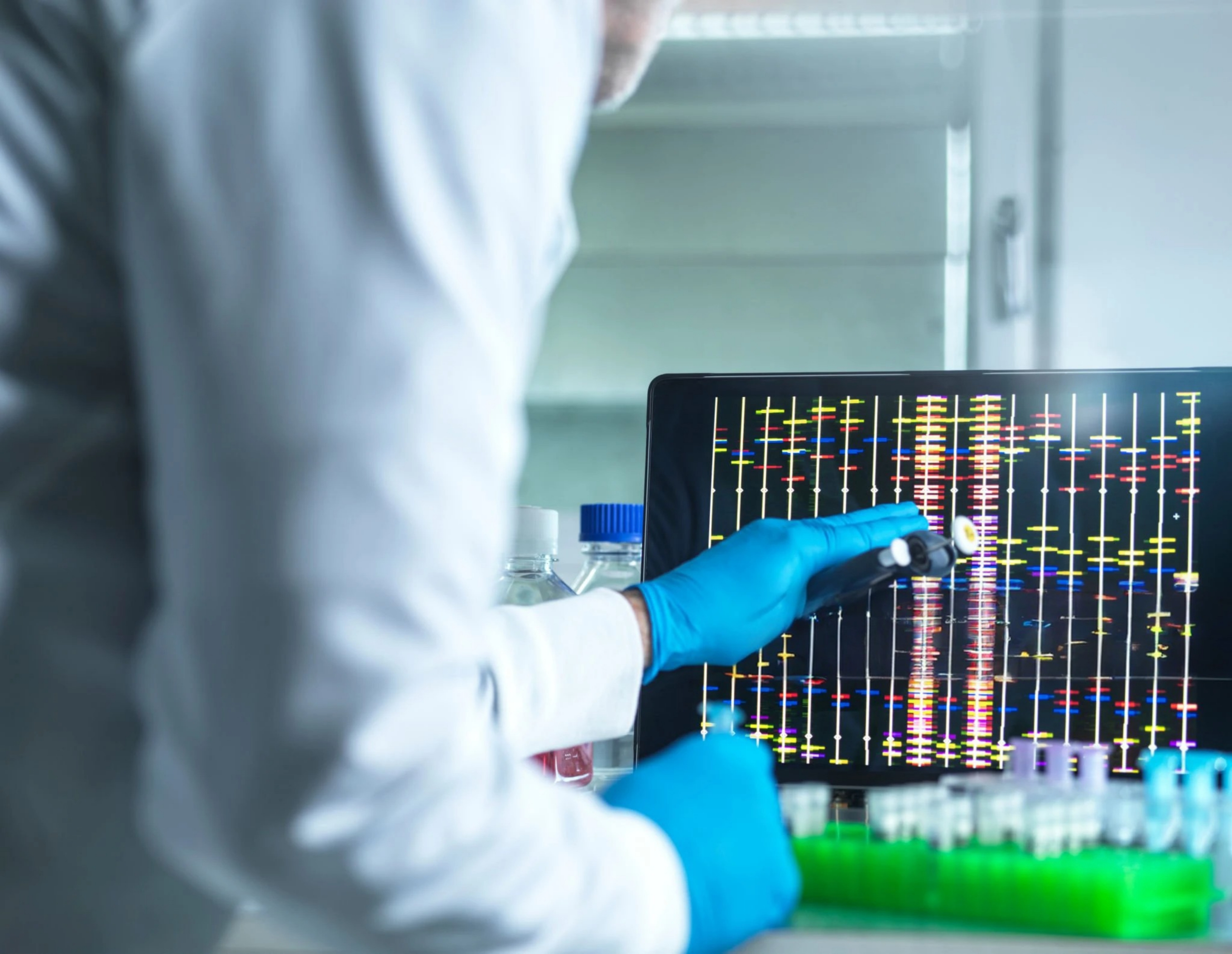 A scientist with rubber gloves points at a computer screen displaying genetic data in a lab setting