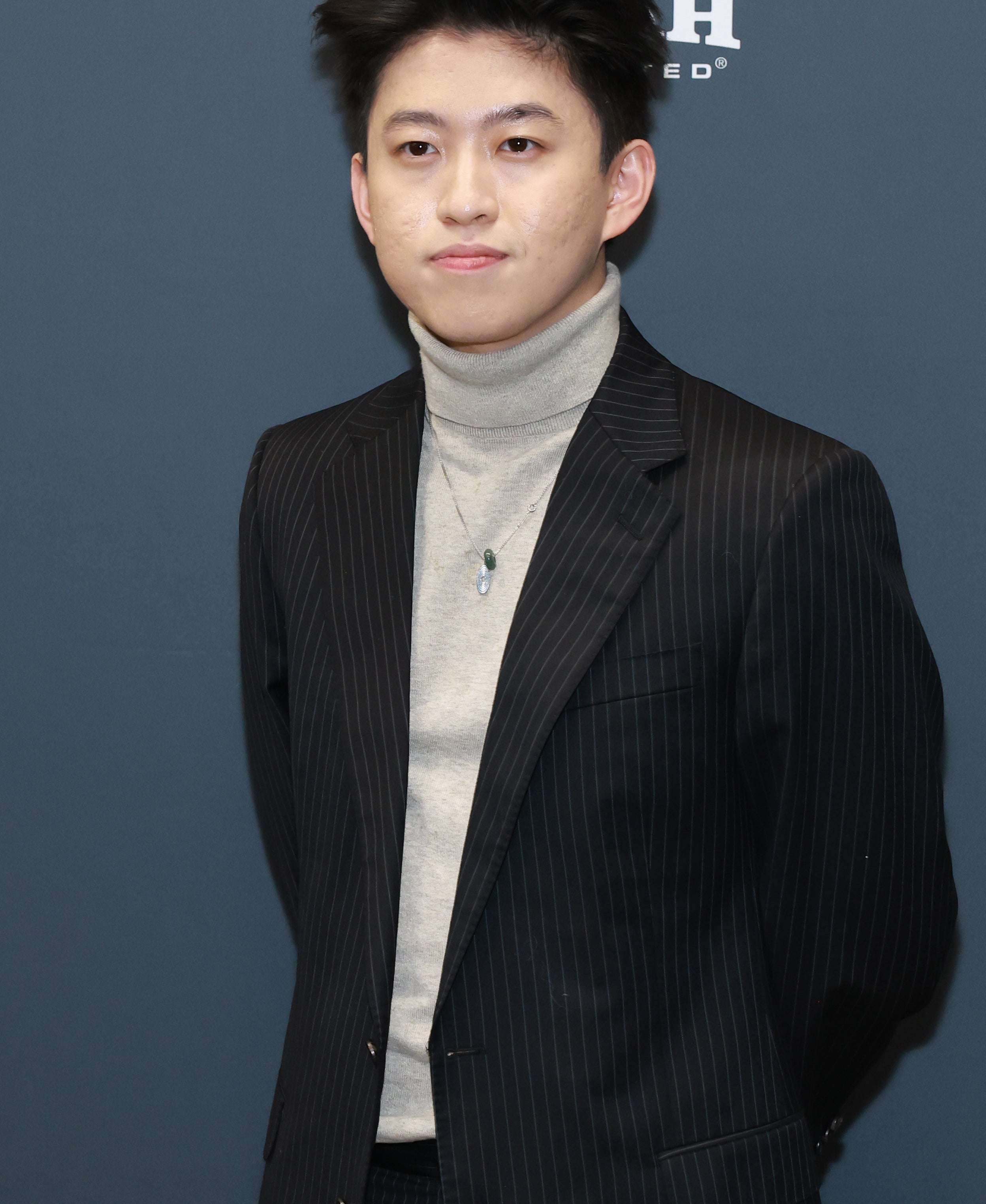 Person in a pinstripe suit and turtleneck stands in front of a blue backdrop, posing for a photo