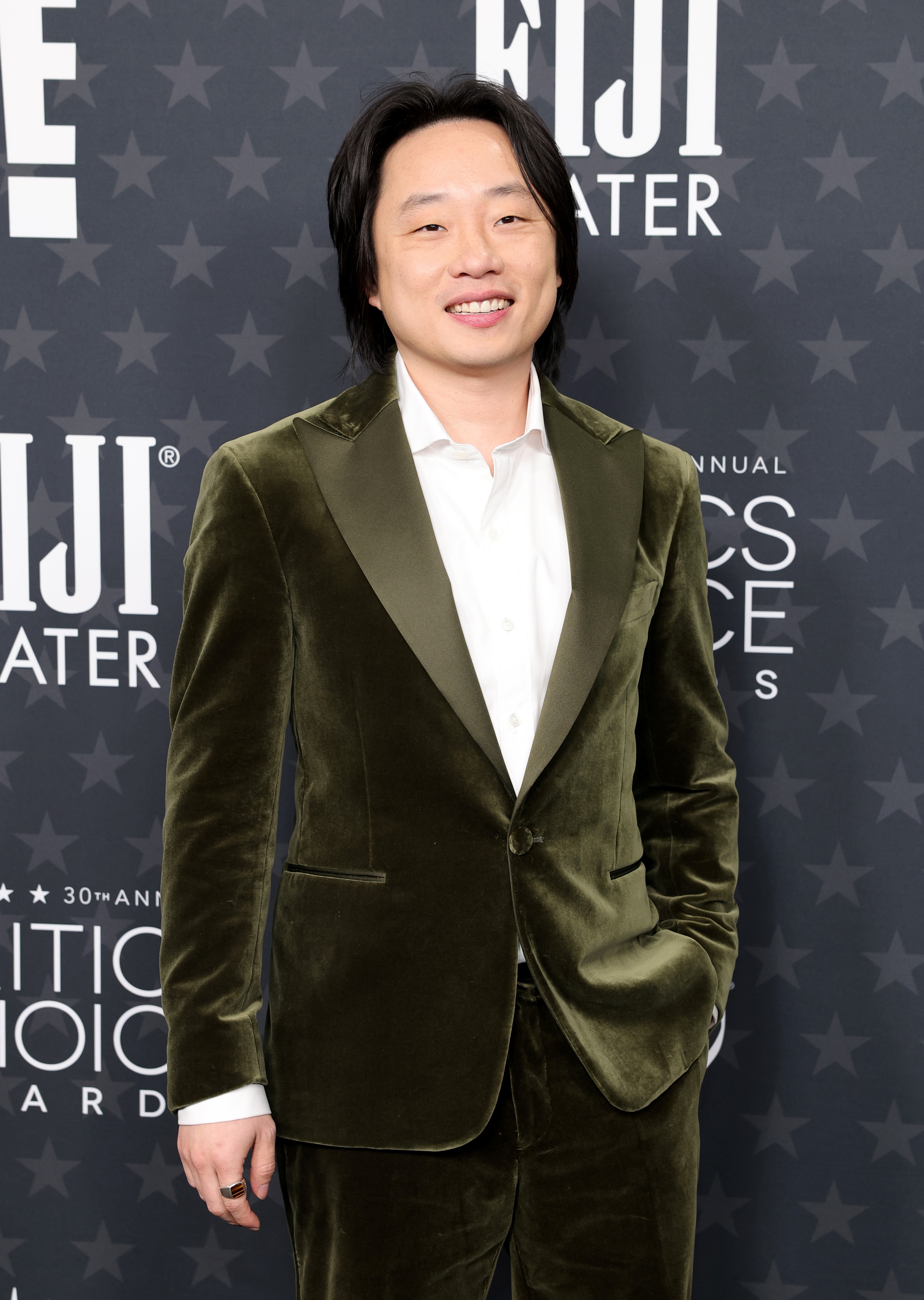 Person in a velvet suit on a red carpet, smiling, with hands in pockets, at an event with branding in the background