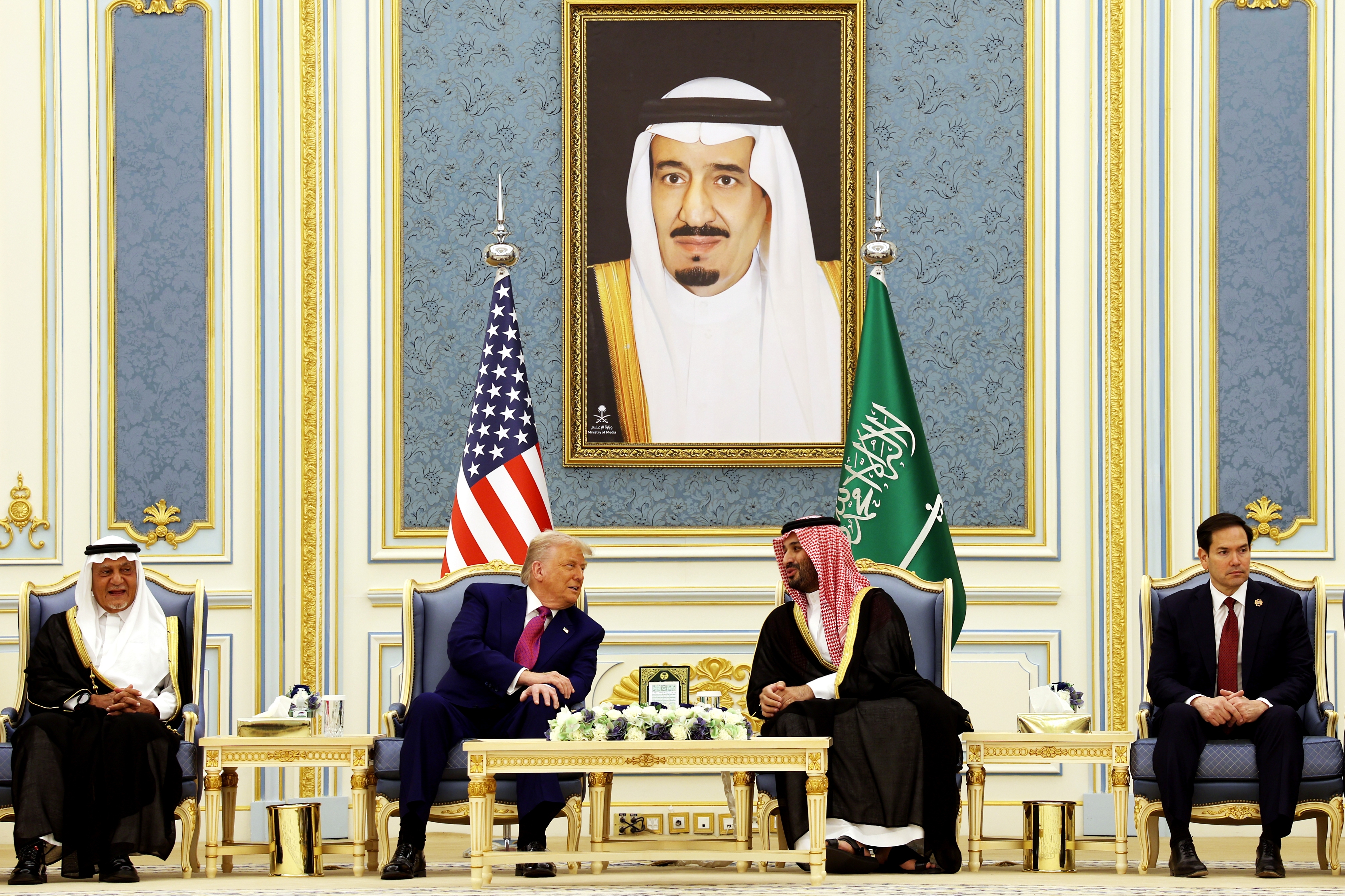 Five men are seated below large portrait in an ornate room. Two men in the center are engaged in conversation, surrounded by American and Saudi flags