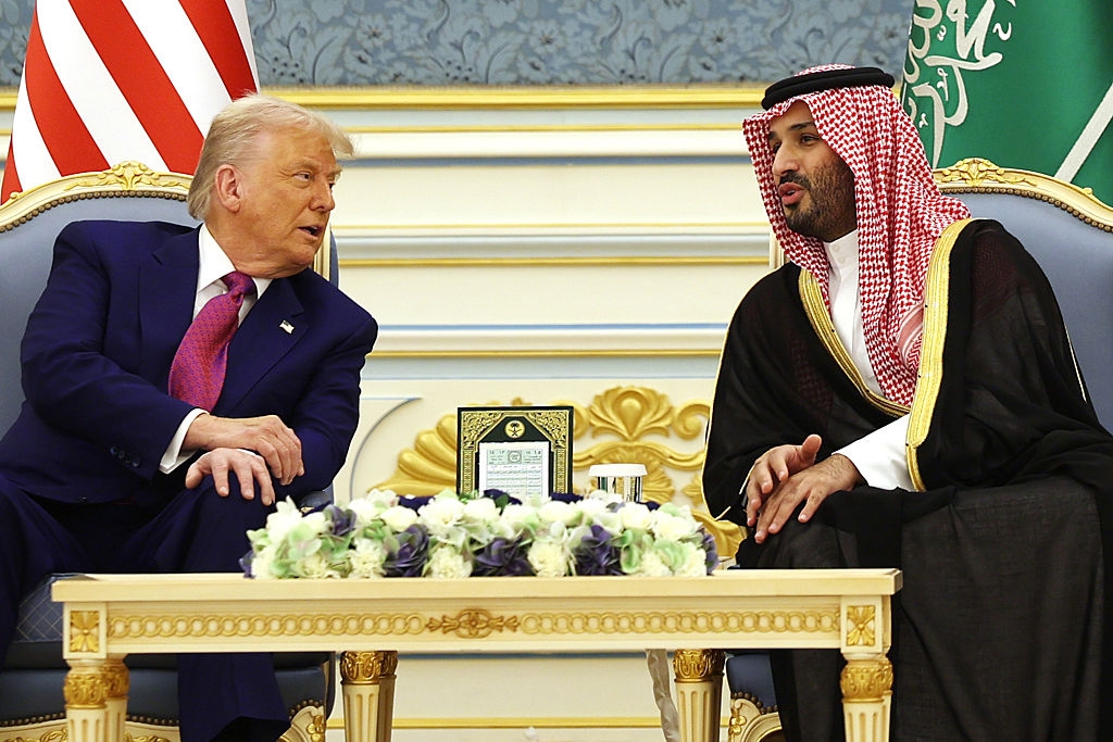 Two men sit and talk in an opulent setting, featuring U.S. and Saudi flags. One wears a suit, and the other is in traditional Saudi attire with a keffiyeh