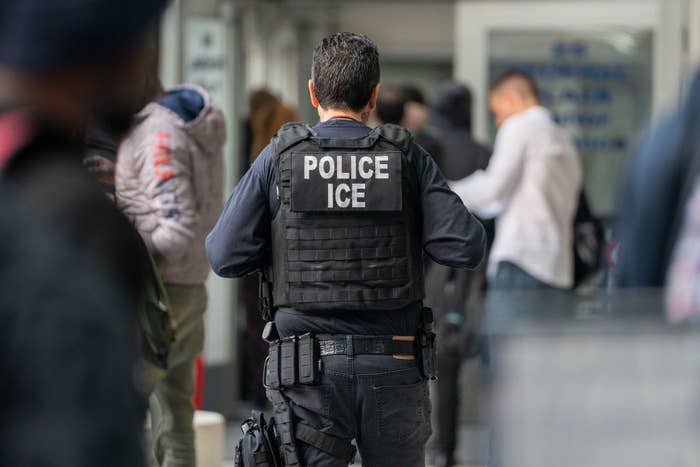 ICE officer in tactical gear faces away, standing in a bustling indoor setting with people nearby