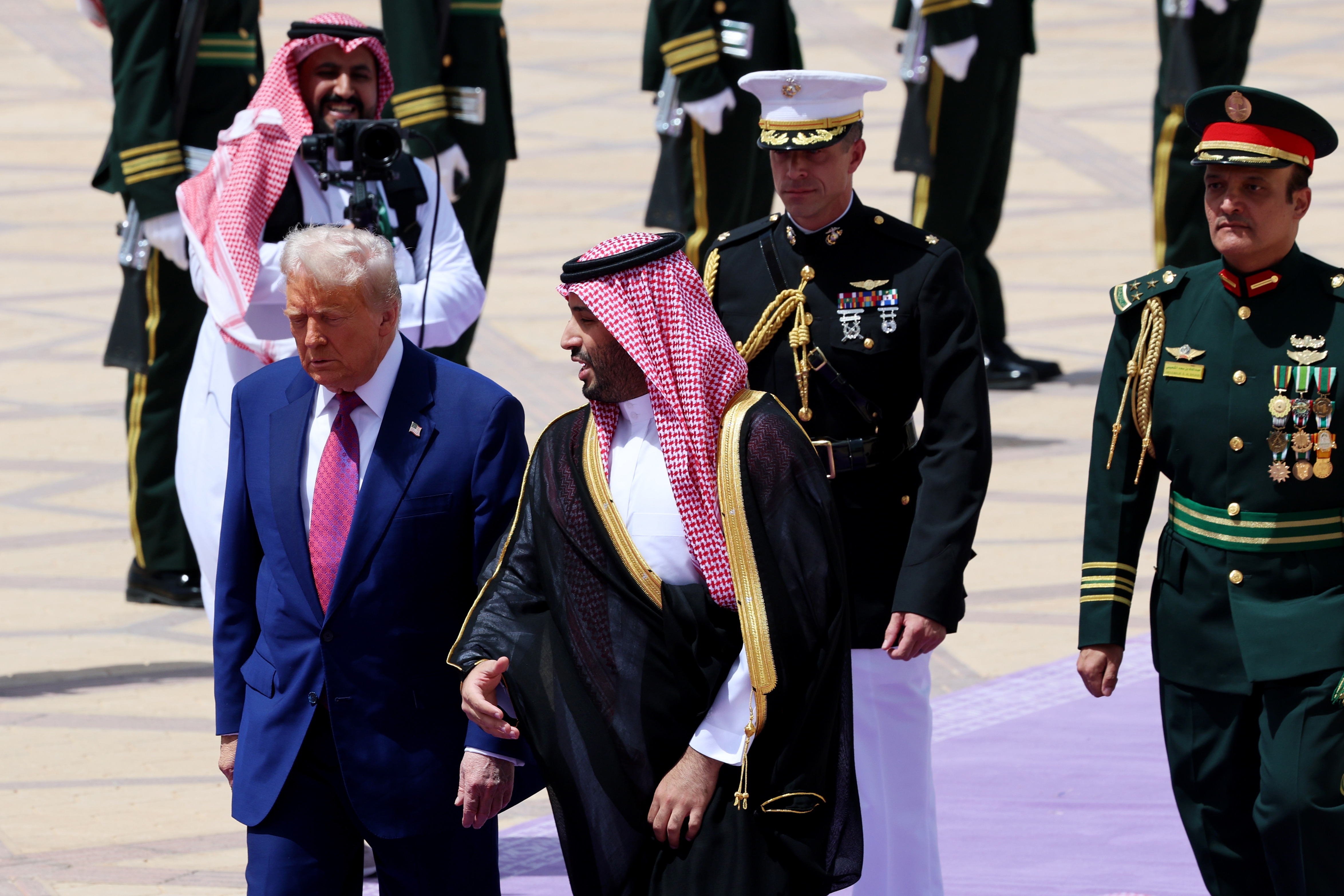 A public figure walking with a dignitary in traditional attire, surrounded by military personnel in a formal setting