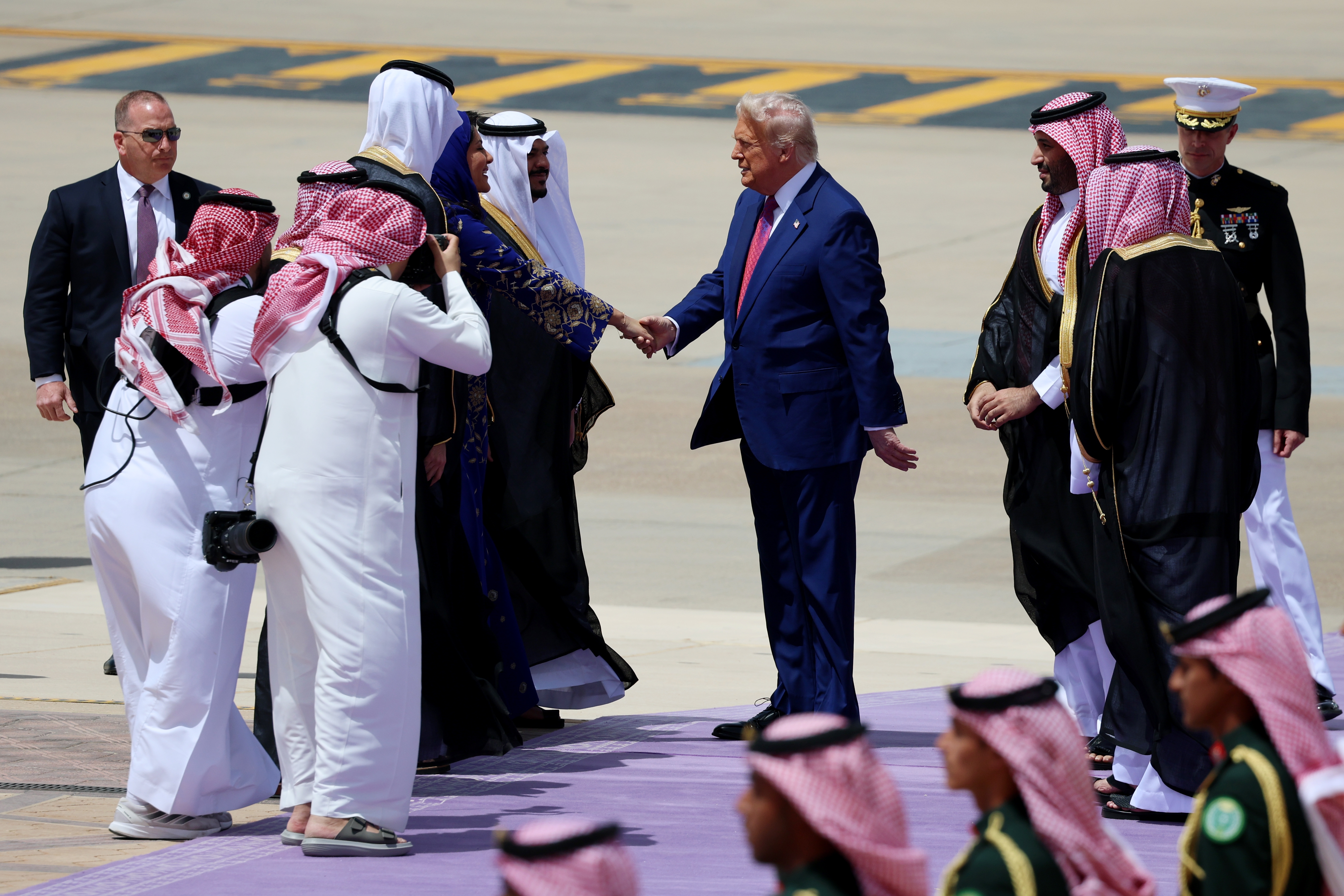 A public figure in a suit shakes hands with officials in traditional attire on a purple carpet, surrounded by photographers and security personnel