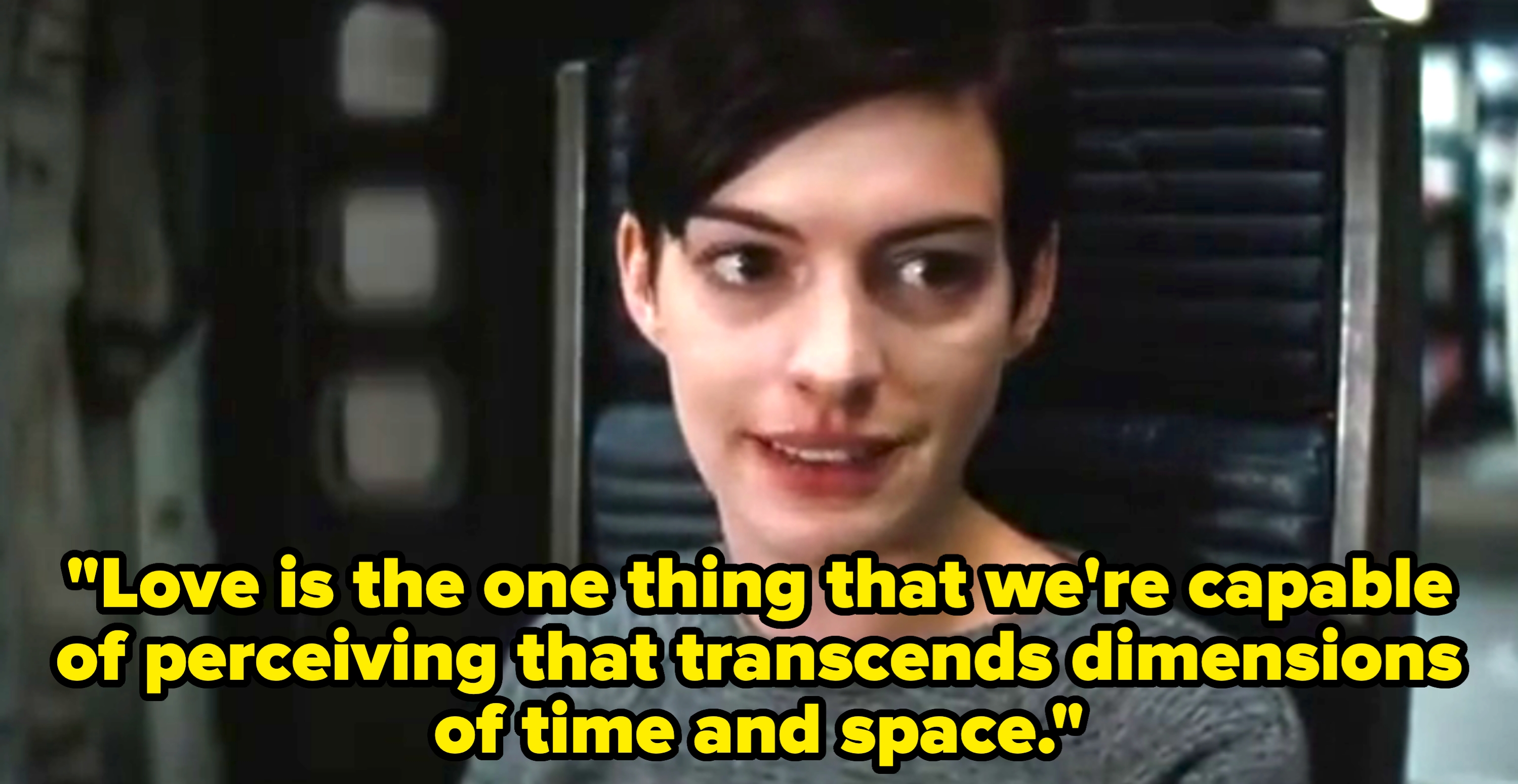 Person in a grey sweater seated in a futuristic setting, speaking or reacting with a slight smile