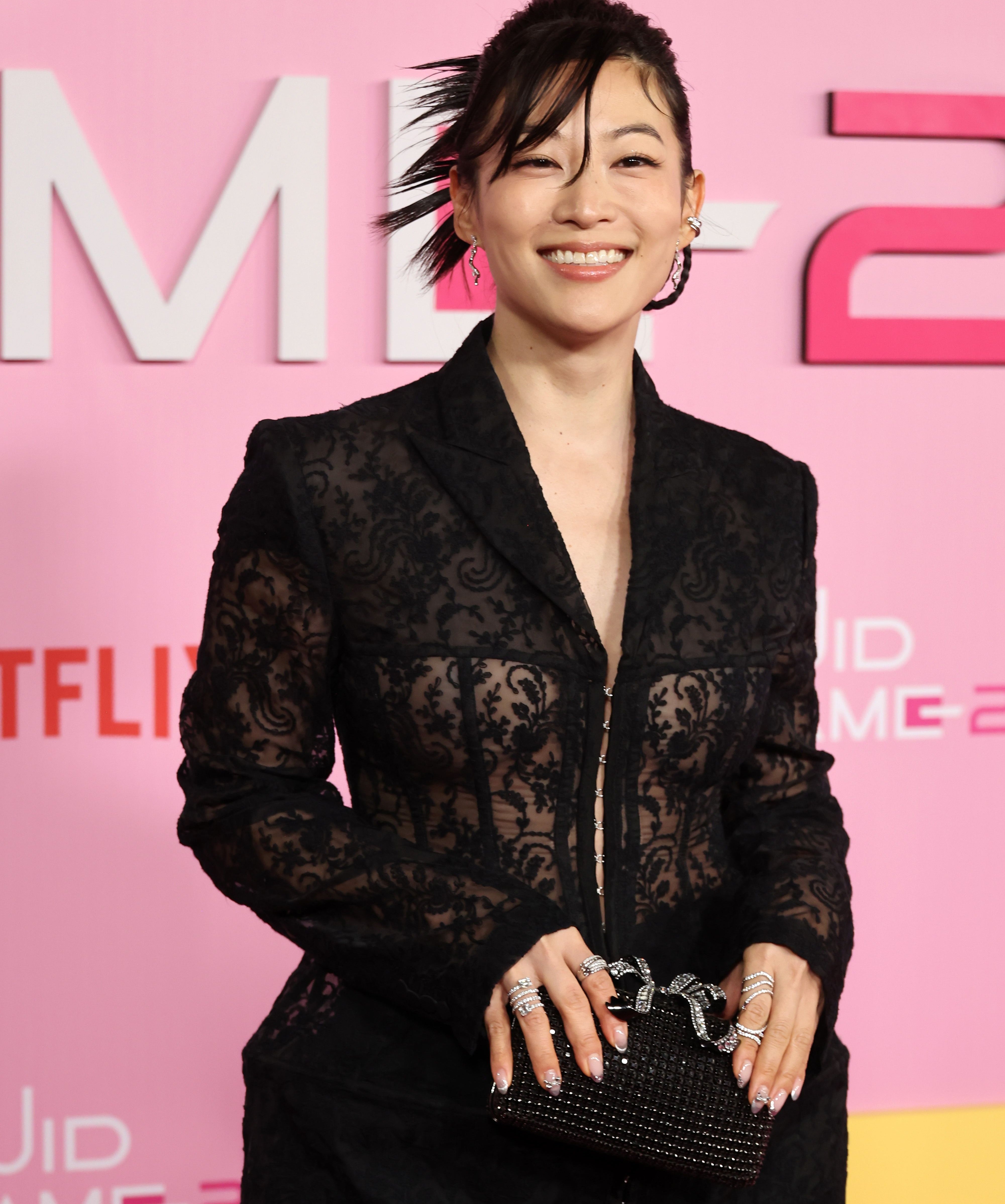 Person in an elegant black lace outfit holds a textured clutch at a formal event, smiling