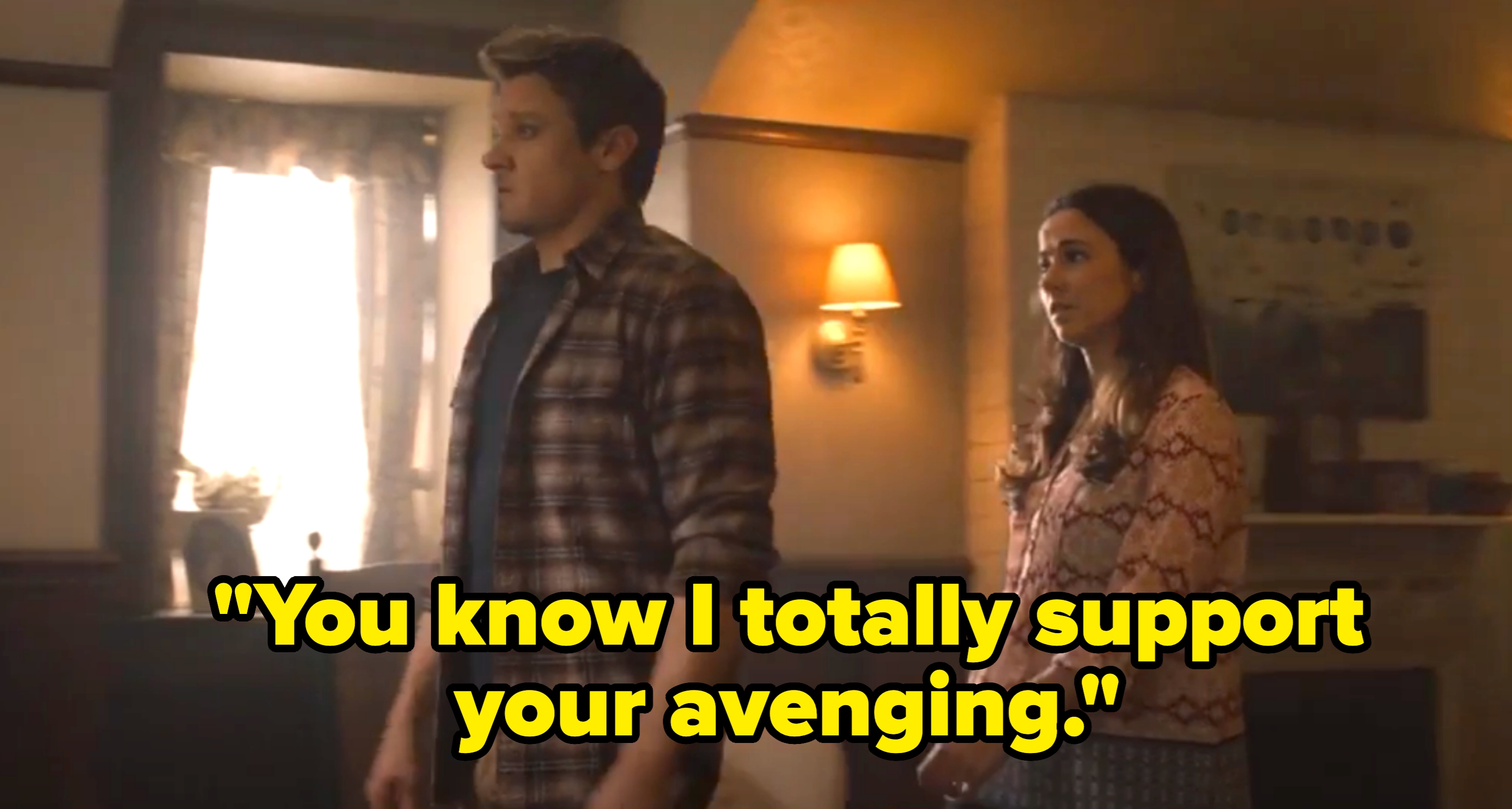 A man in a plaid shirt and a woman in a patterned top stand in a dimly lit room, appearing thoughtful