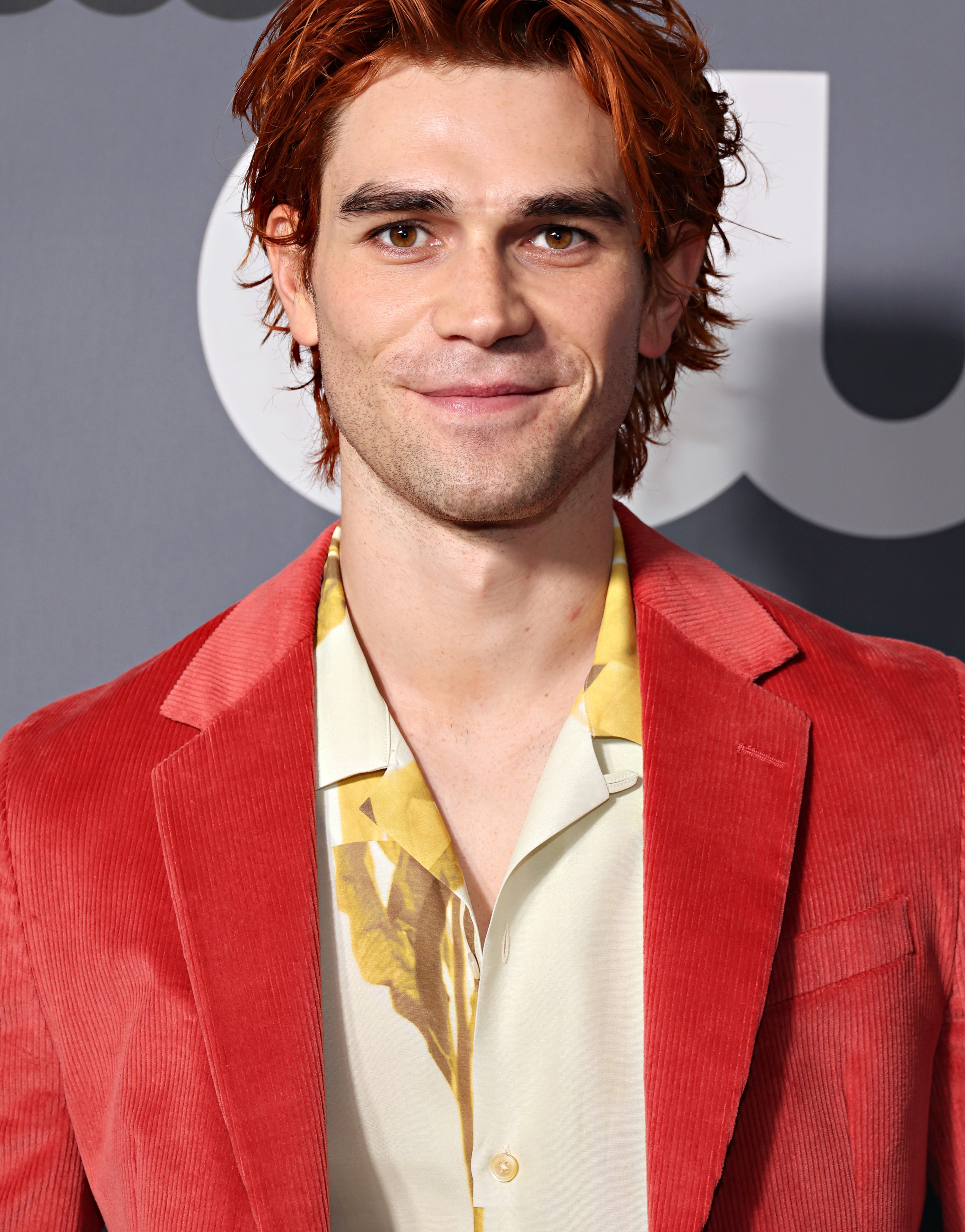 Person in a red suit and floral shirt, smiling at an event backdrop