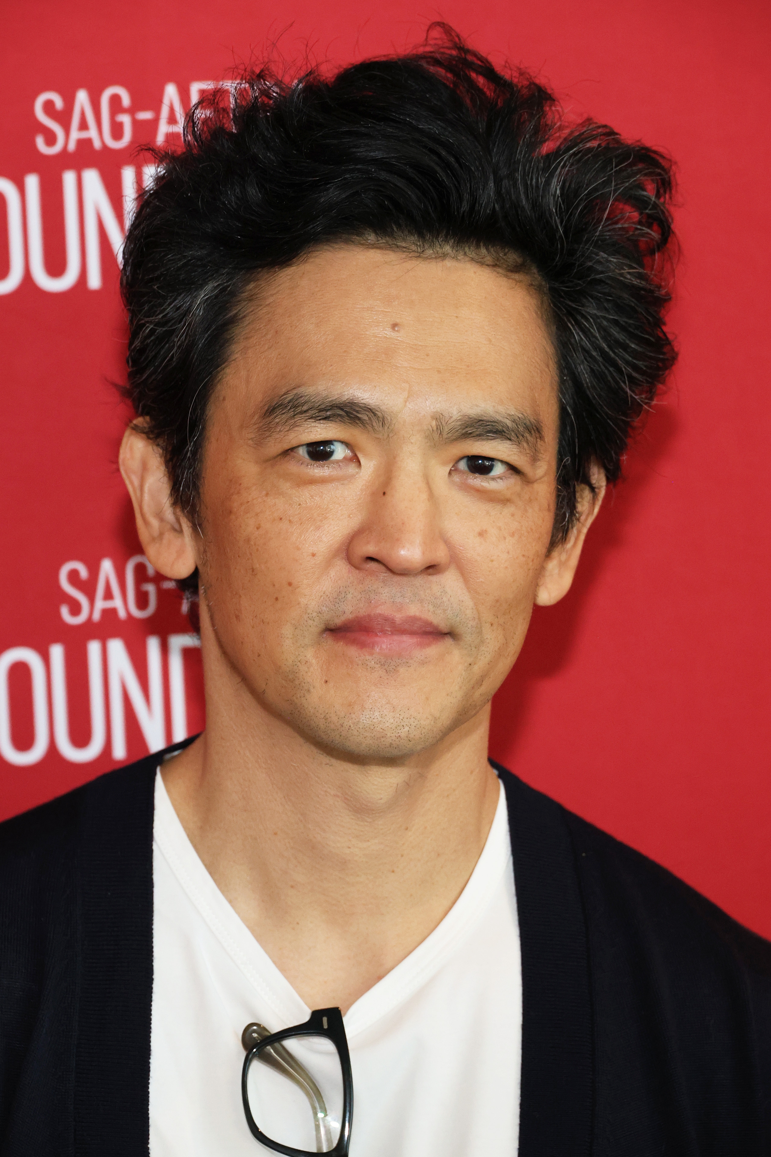 Person stands in front of a backdrop with event lettering, wearing a white shirt and dark jacket, holding glasses