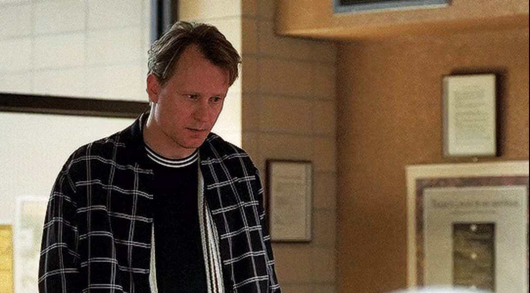 Person in a checkered shirt looks pensively at someone off-screen in a casual setting