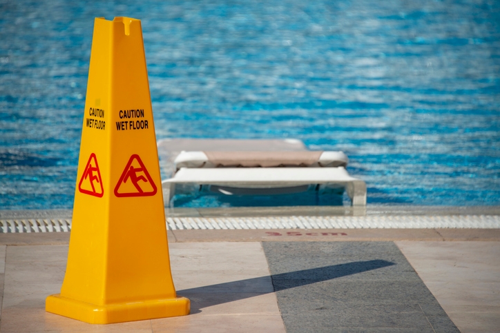 Yellow &quot;Caution Wet Floor&quot; sign by a swimming pool, casting a shadow on the tiled ground