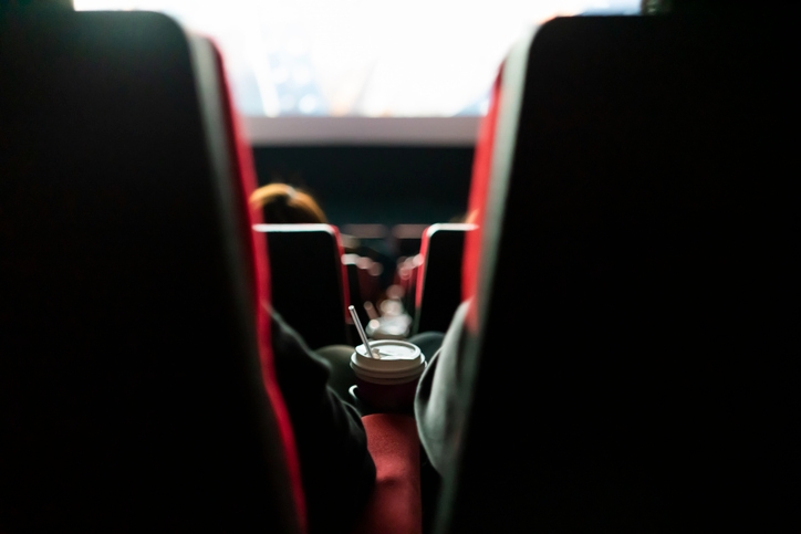 View from behind cinema seats, focusing on a drink with a straw held by a person, facing a bright movie screen ahead