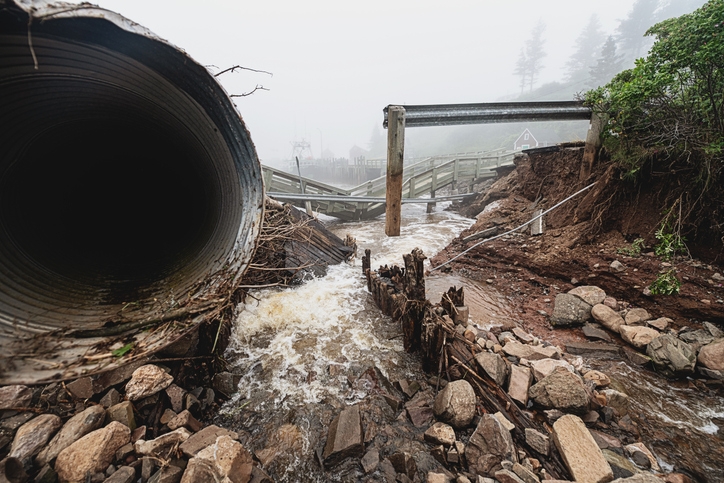 Collapsed bridge over a rushing stream with visible erosion and large drainage pipe nearby. Rocks and debris surround the scene, highlighting flood damage
