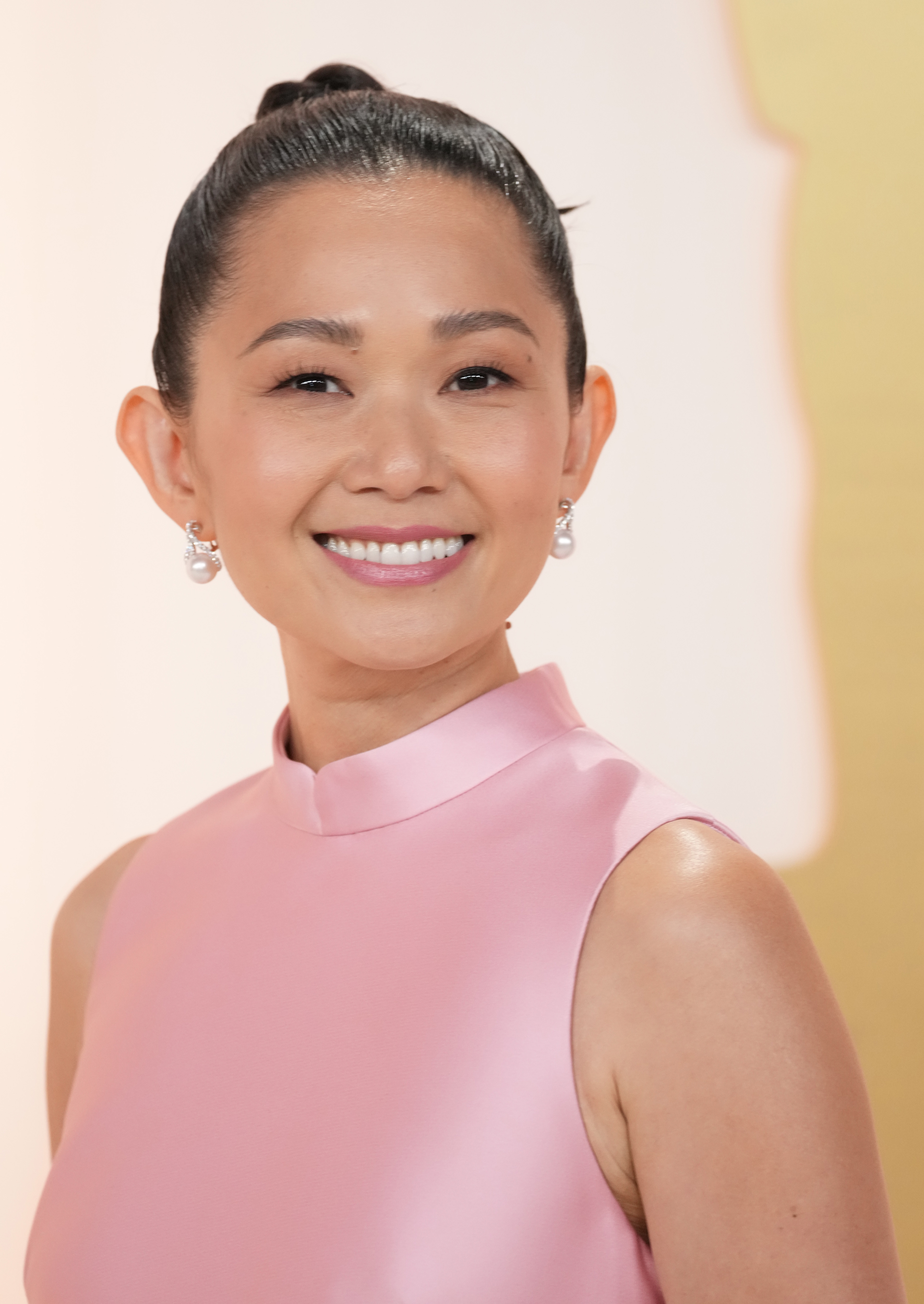 Hong Chau smiling in a sleek, high-neck pink gown with pearl earrings, hair in a neat bun
