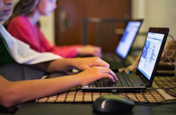Two people focus intently on laptops on a table, with hands typing; blurred background suggests a home setting